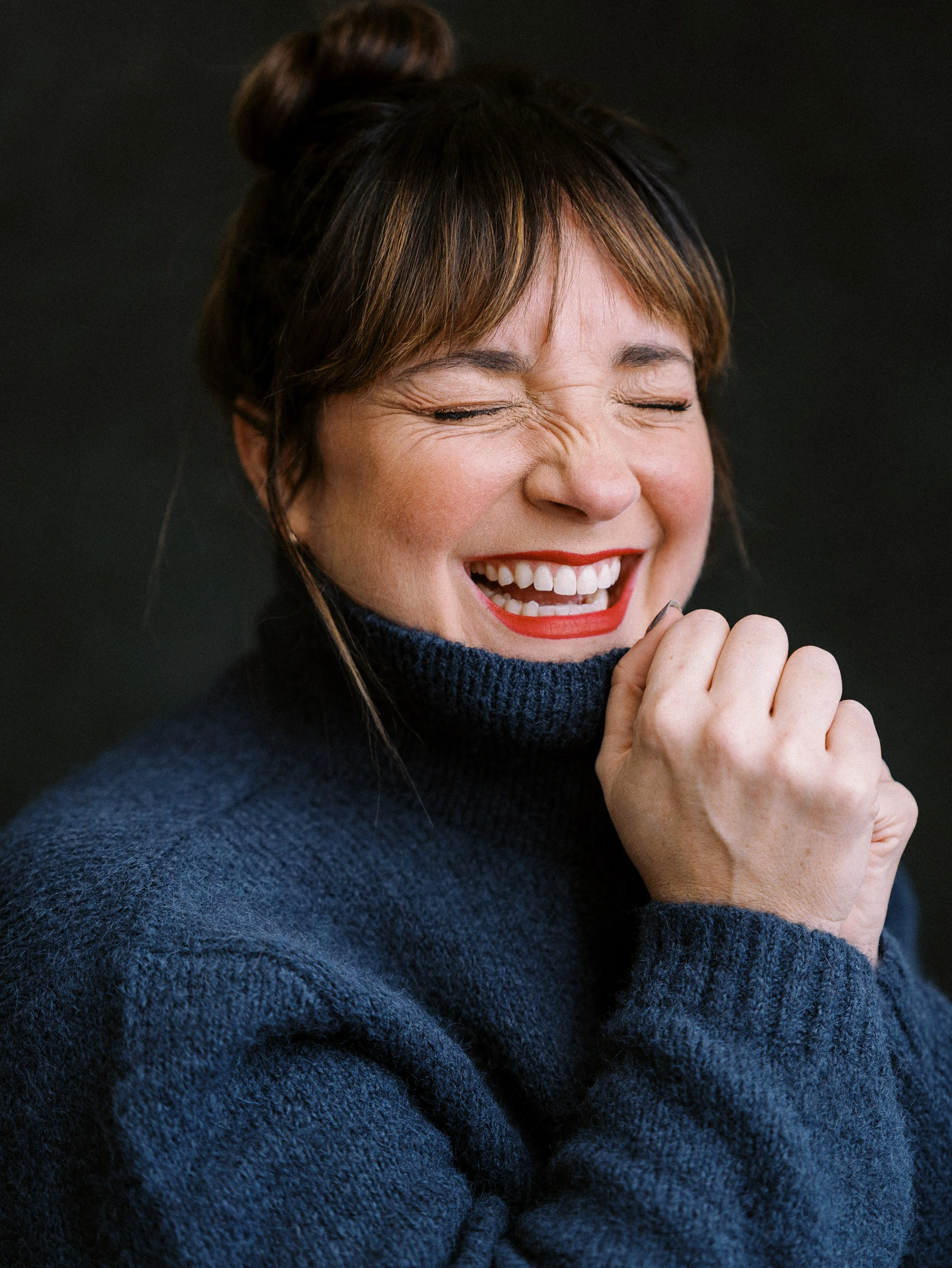 A woman with brown hair styled in a bun, wearing a dark blue sweater, laughing with eyes closed and hand near her face, against a black background.