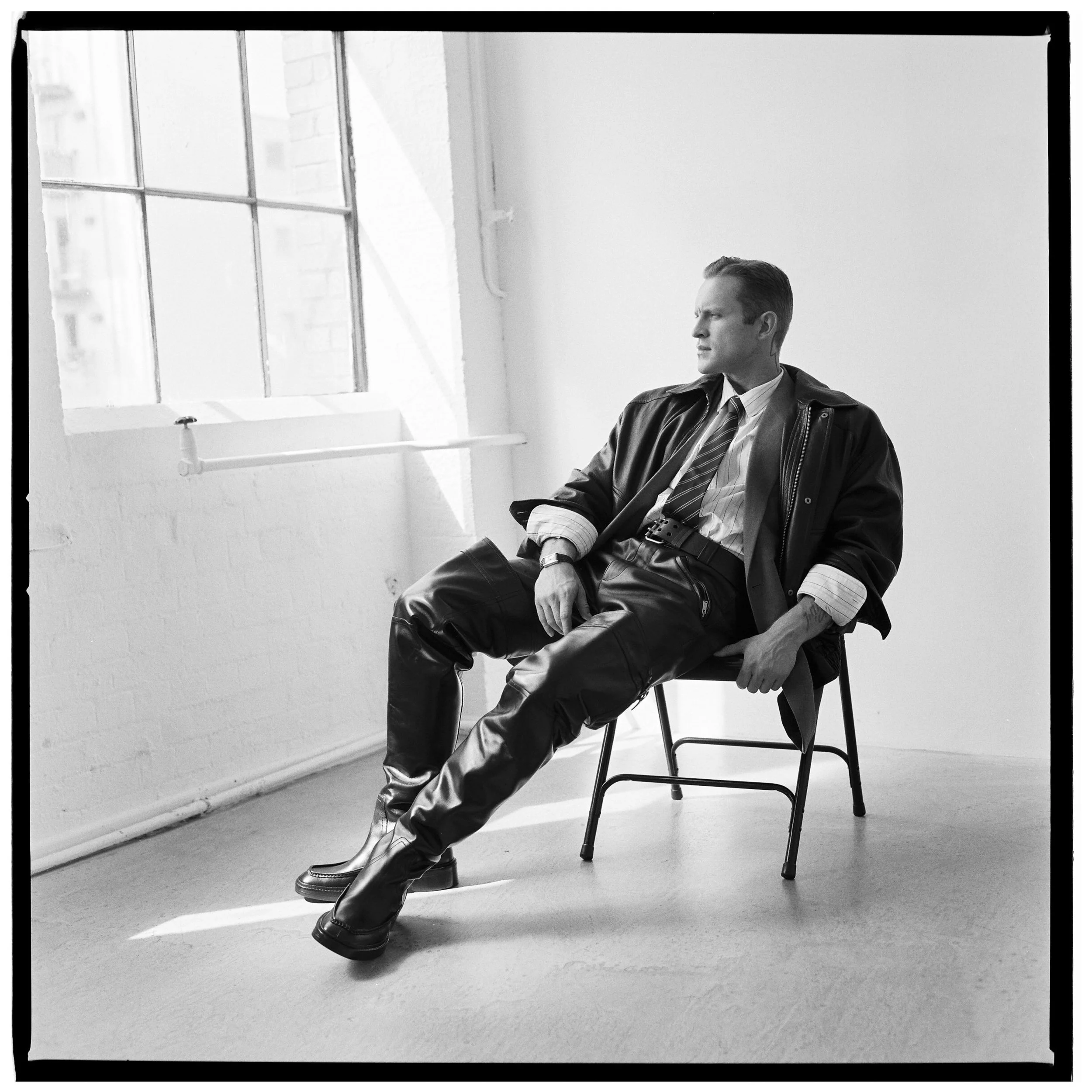 A black and white photo of a man sitting on a chair in a minimalistic room with a large window, wearing a leather jacket, dress shirt, tie, and leather pants, looking to the side with a serious expression.