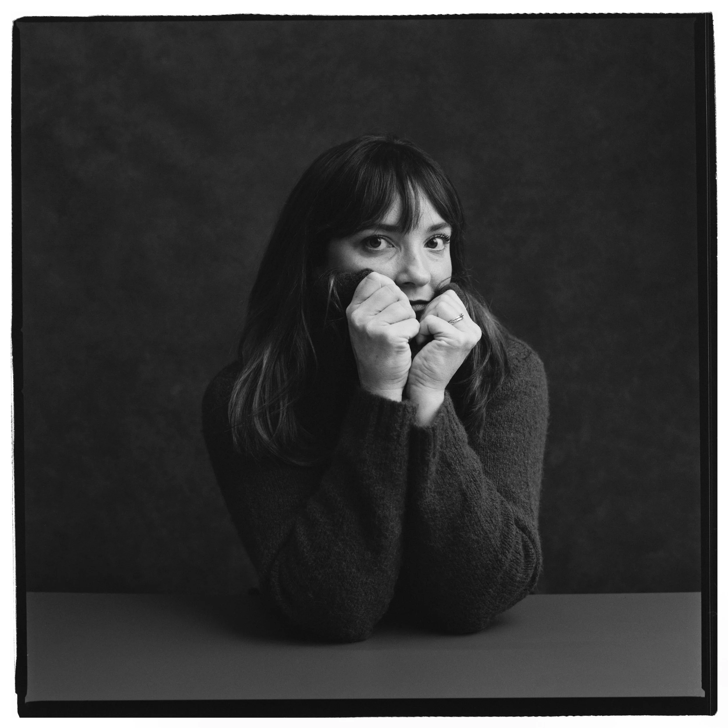 Black and white portrait of a woman with dark hair and bangs, resting her face on her fists, wearing a dark sweater, looking directly at the camera with a contemplative expression.