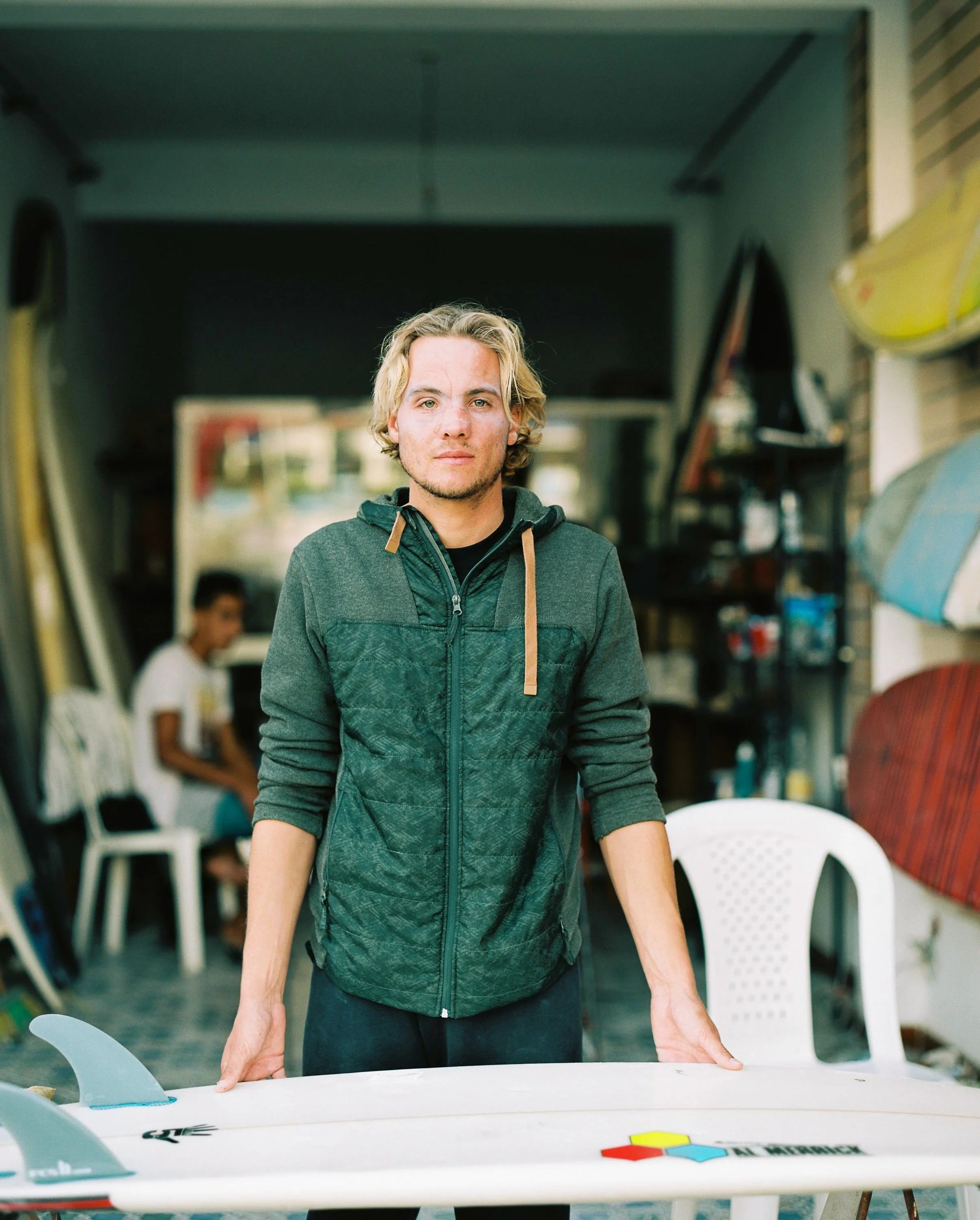 A young man with blond hair and a green jacket standing behind a table with a surfboard on it in a surf shop.