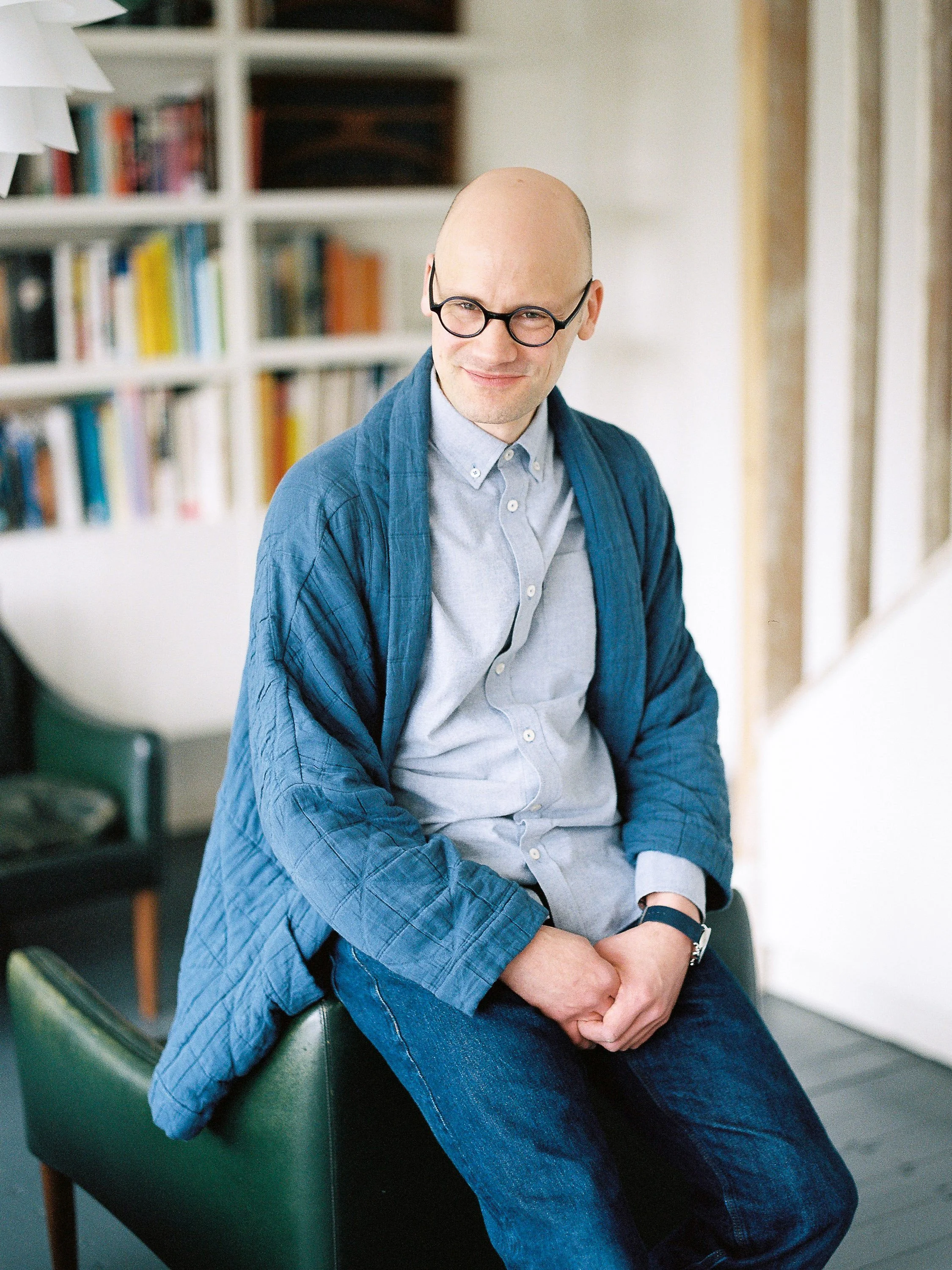 A bald man wearing glasses, a light blue button-down shirt, a dark blue quilted jacket, and jeans, sitting on a green chair in a room with bookshelves and a white wall.