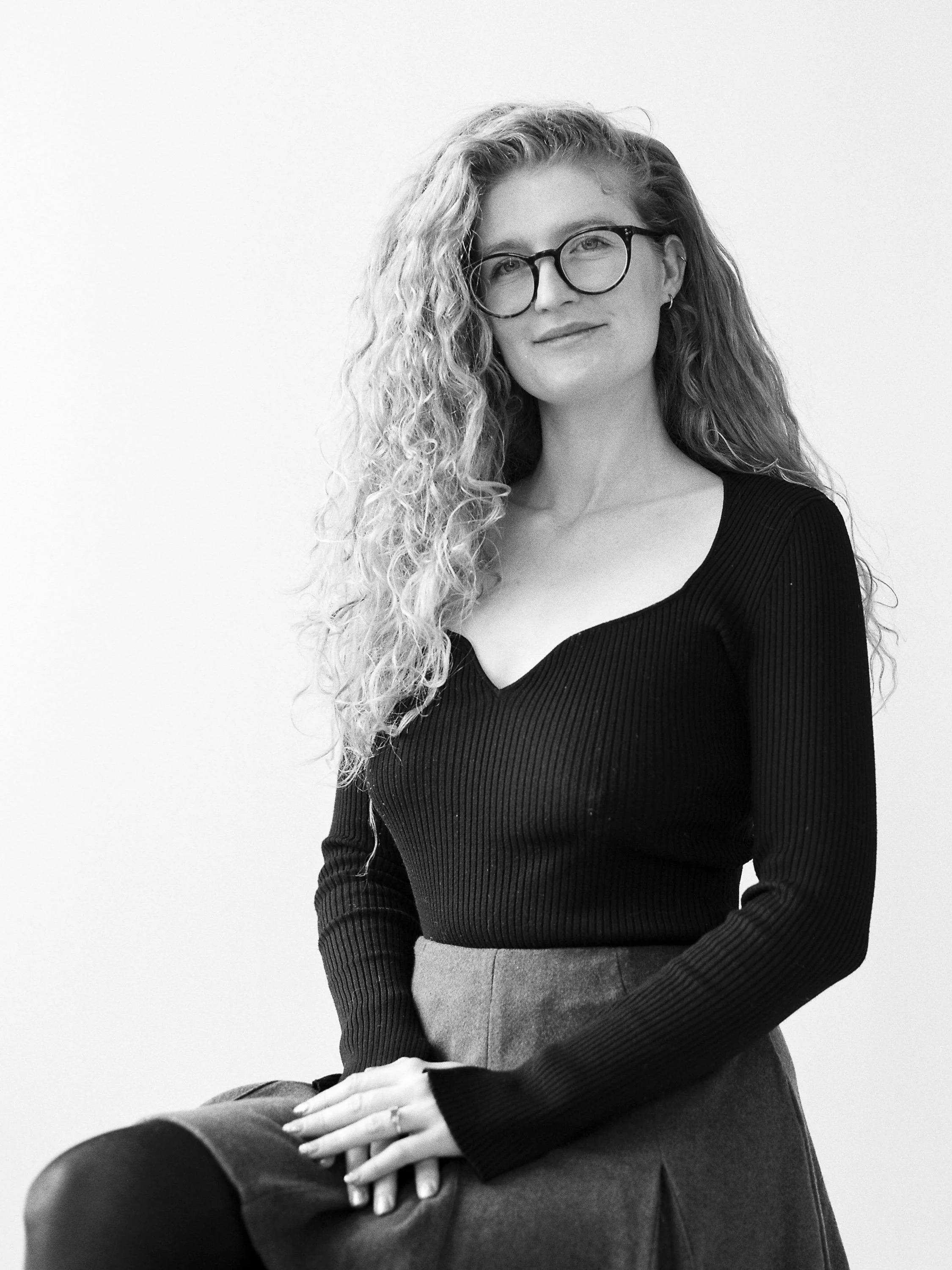 A black and white photo of a woman with long curly hair and glasses, sitting with one hand resting on her lap, wearing a black top and a skirt, looking at the camera.