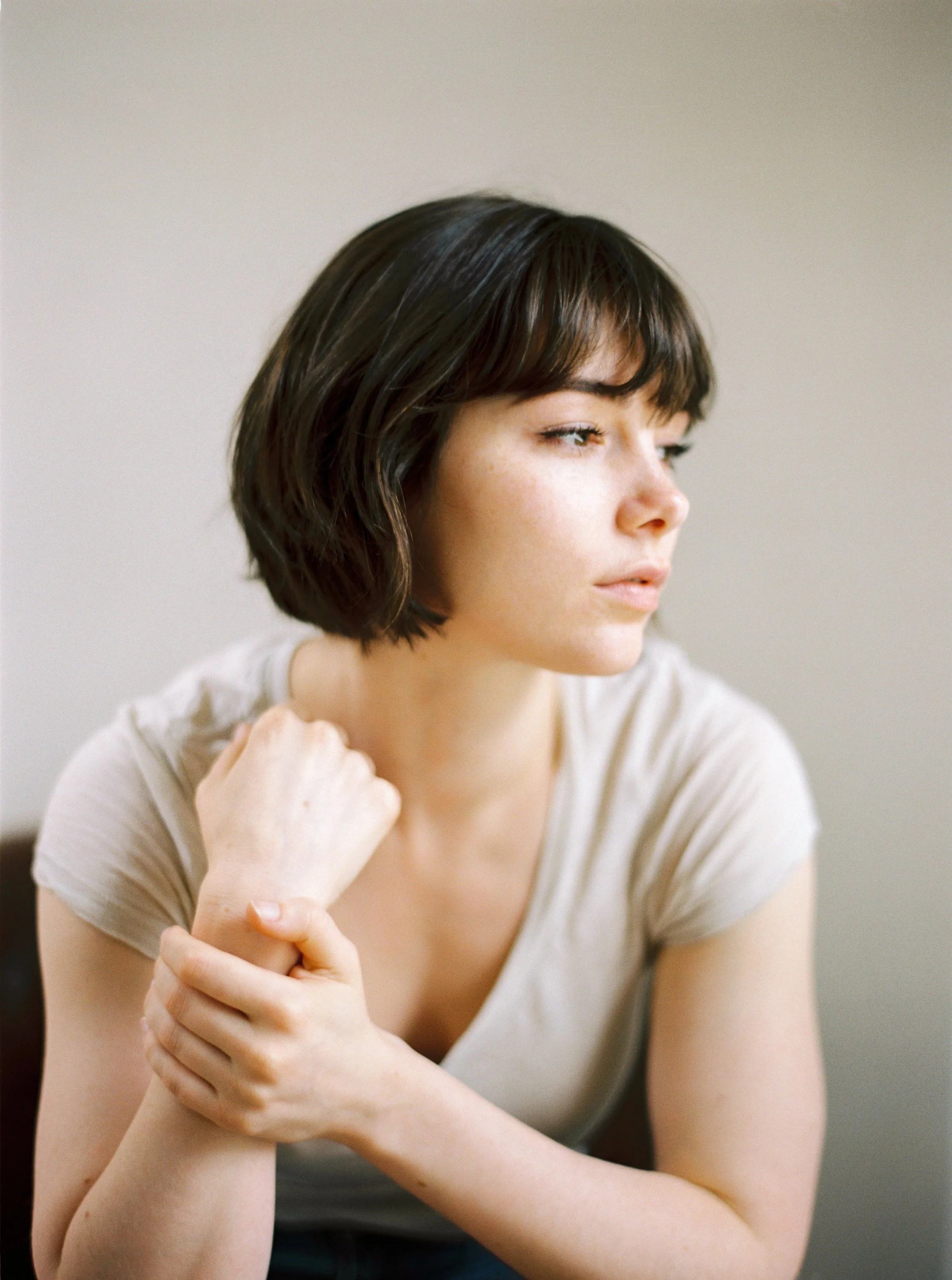 A young woman with short dark hair and fair skin sits with her left arm across her chest and her right hand holding her left wrist. She gazes thoughtfully to the right with a neutral expression, wearing a light-colored T-shirt in a bright, softly lit