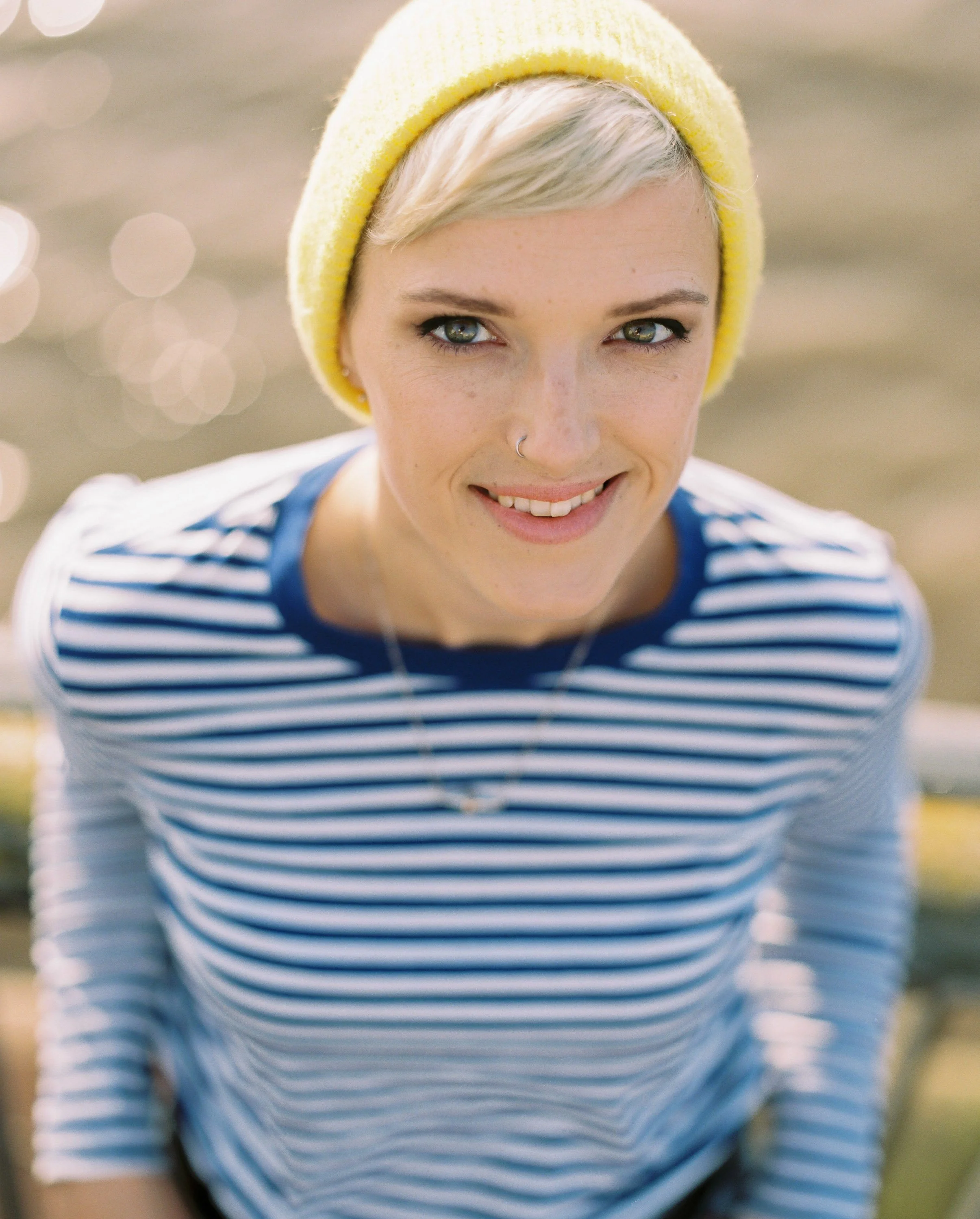 A young woman with short blonde hair, wearing a yellow knit beanie and a blue and white striped shirt, smiling outdoors with water in the background.
