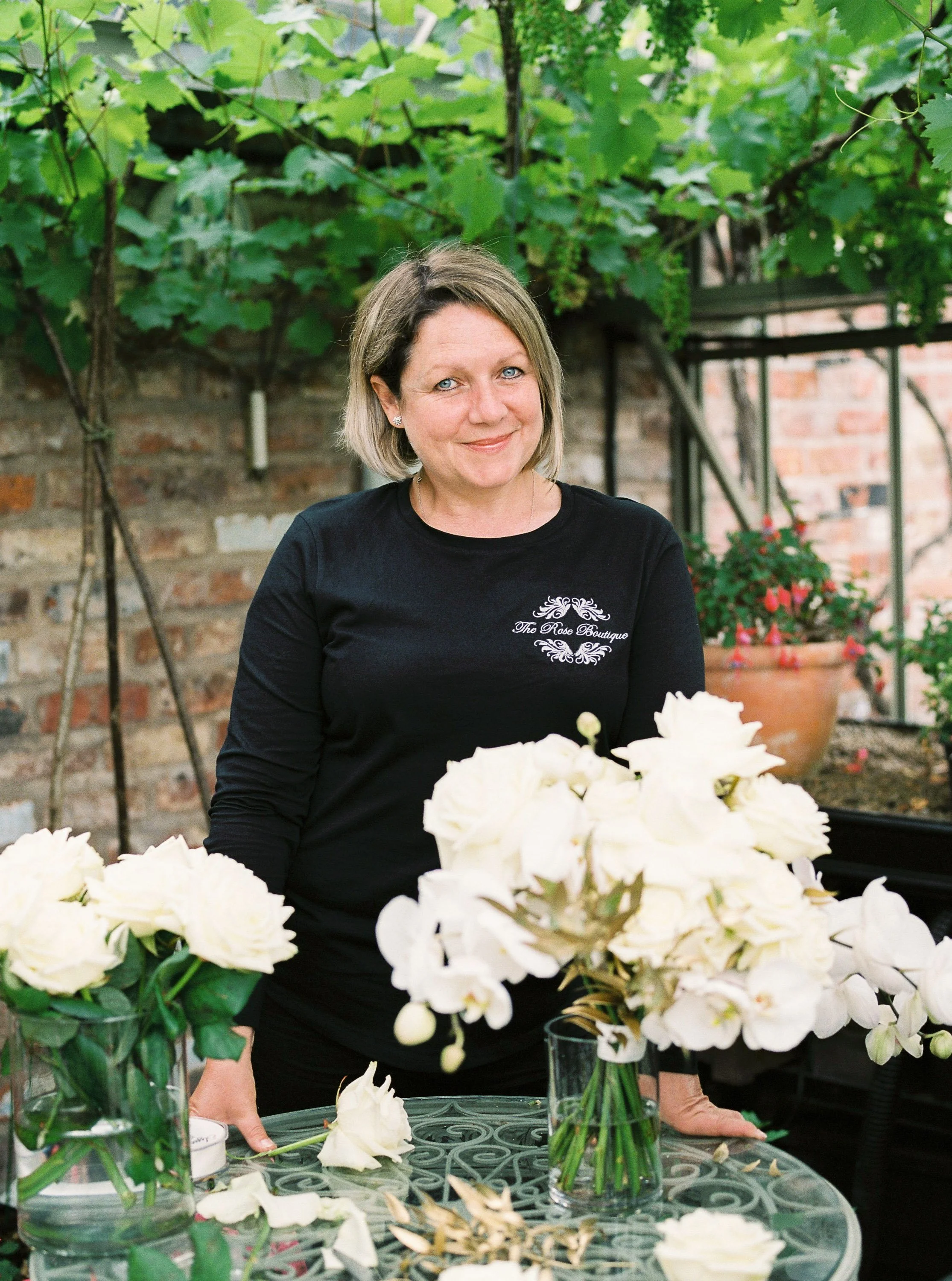 A woman with blonde hair and wearing a black long-sleeve shirt that has 'The Rose Boutique' embroidered on it, standing behind a glass table with white rose flowers in glass vases, in a greenhouse or garden setting with vine-covered brick walls in th