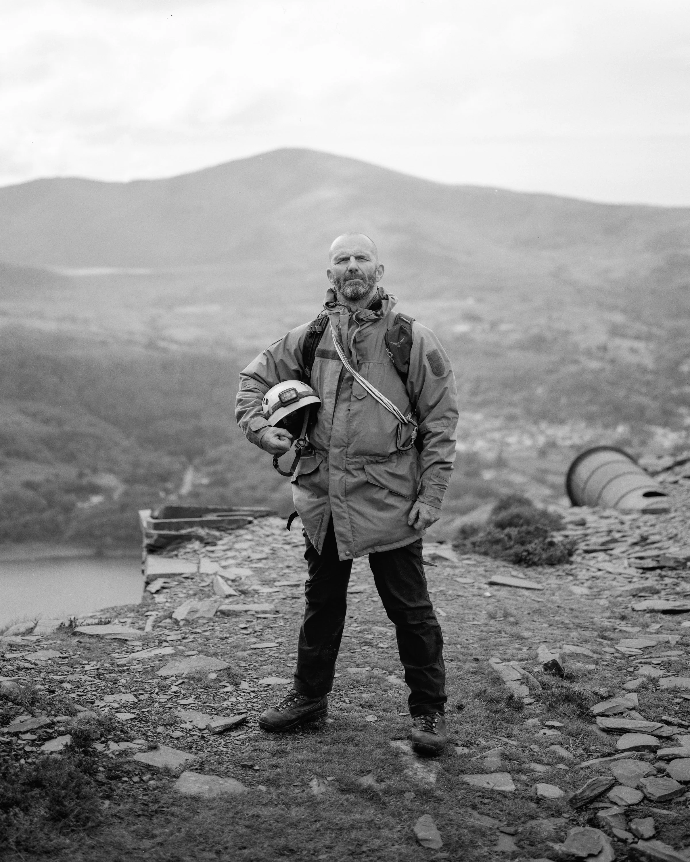 A man standing on a rocky mountain trail with a helmet in his hand, surrounded by scenic mountains and a cloudy sky.