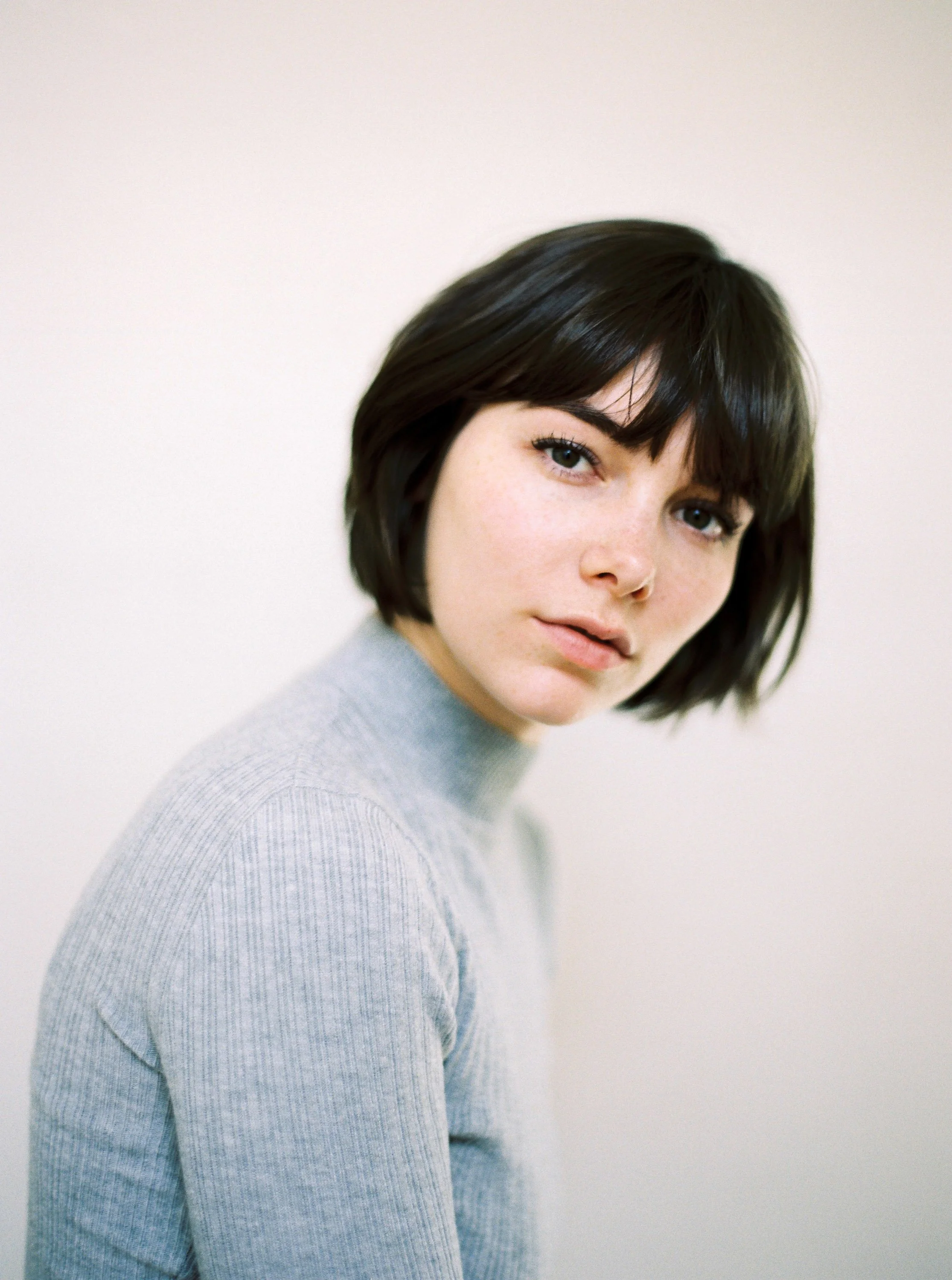 A portrait of a young woman with short dark hair and fair skin, wearing a light gray turtleneck sweater, against a plain light-colored background.