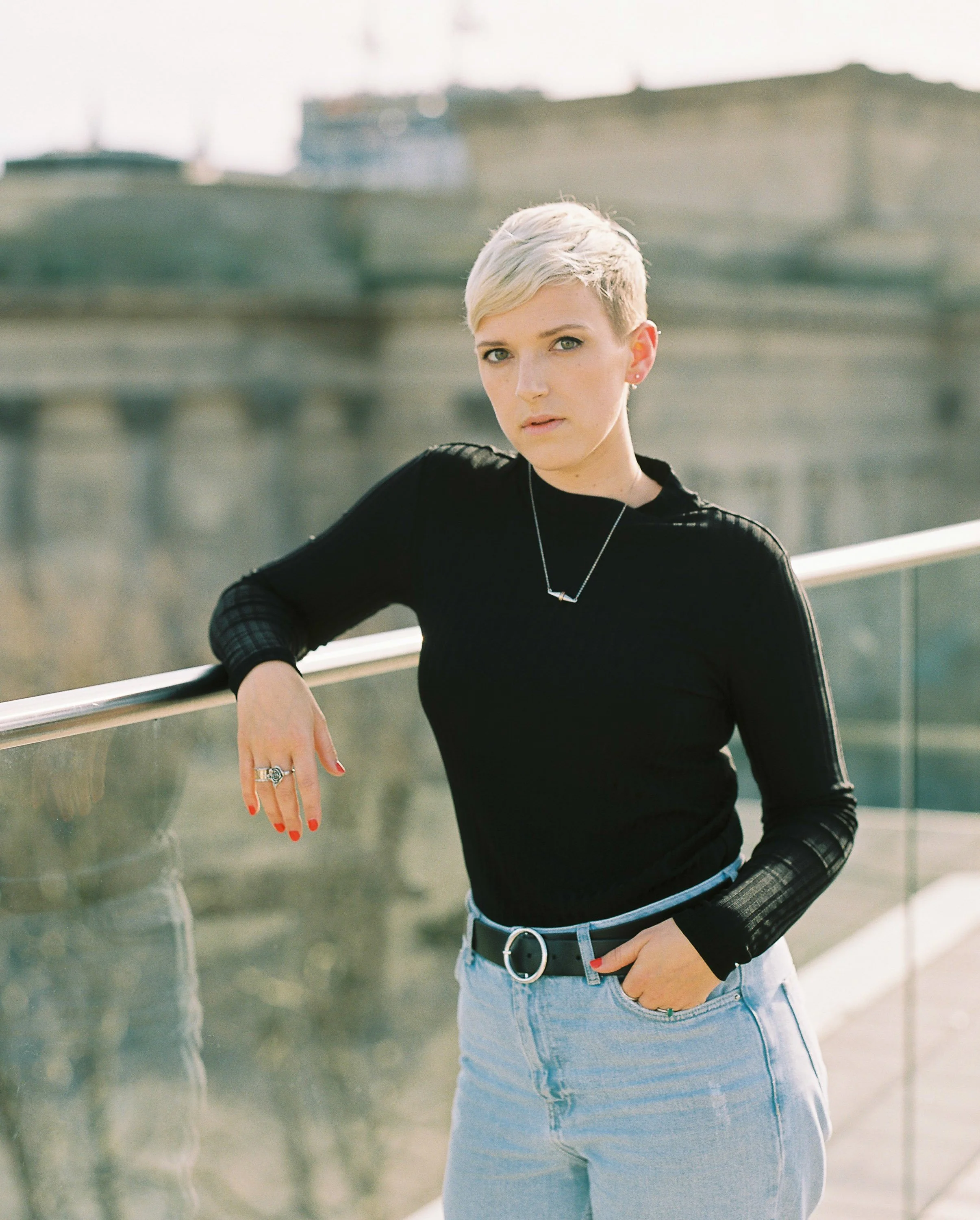 Woman with short blond hair wearing a black long-sleeve top and light blue jeans, standing on a bridge with a cityscape in the background.