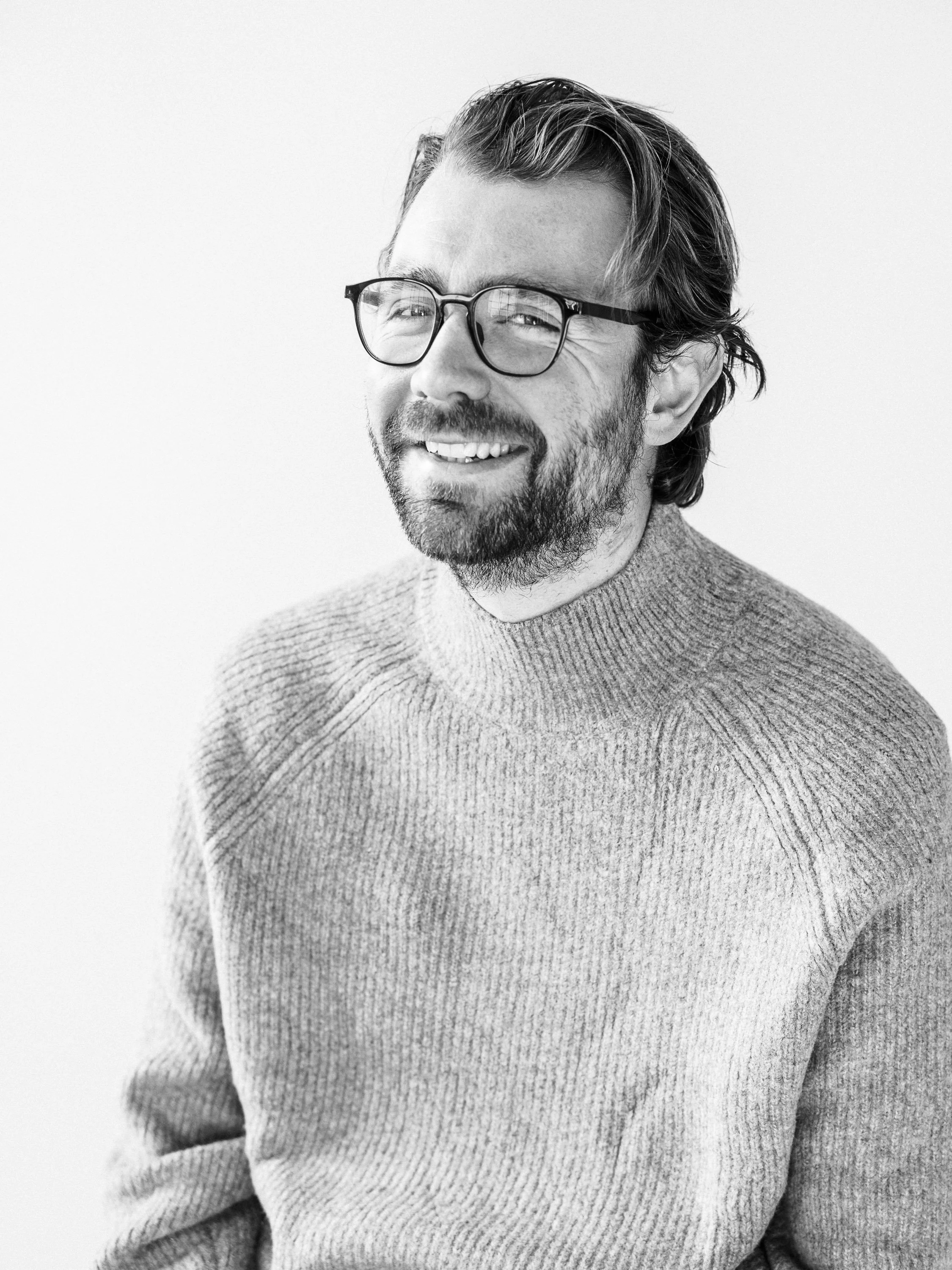 Black and white portrait photo of a middle-aged man with glasses, a beard, and wavy hair, smiling and wearing a knit sweater.