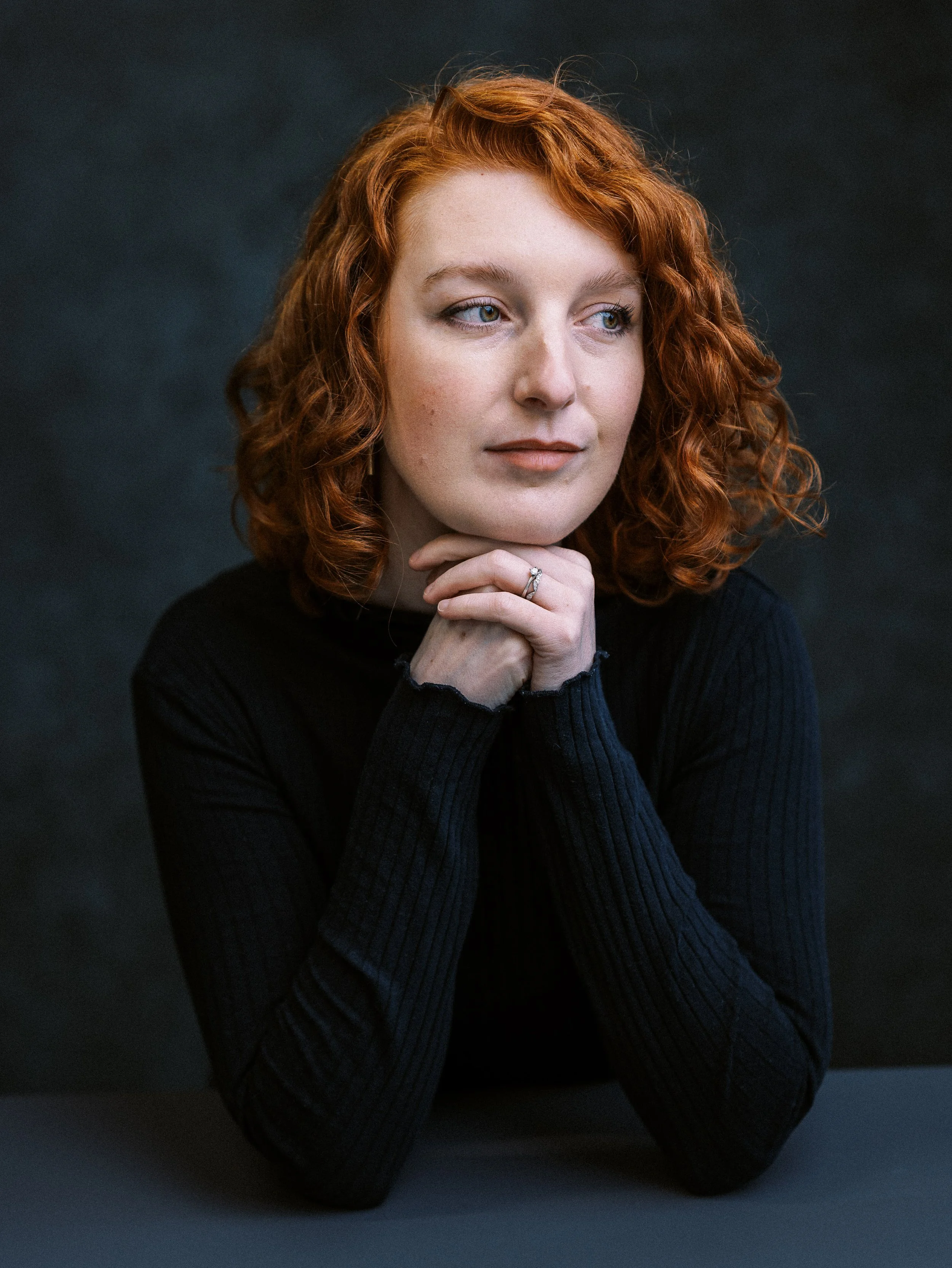 A woman with red curly hair wearing a black long-sleeve shirt, resting her chin on her clasped hands, looking thoughtfully to the side.