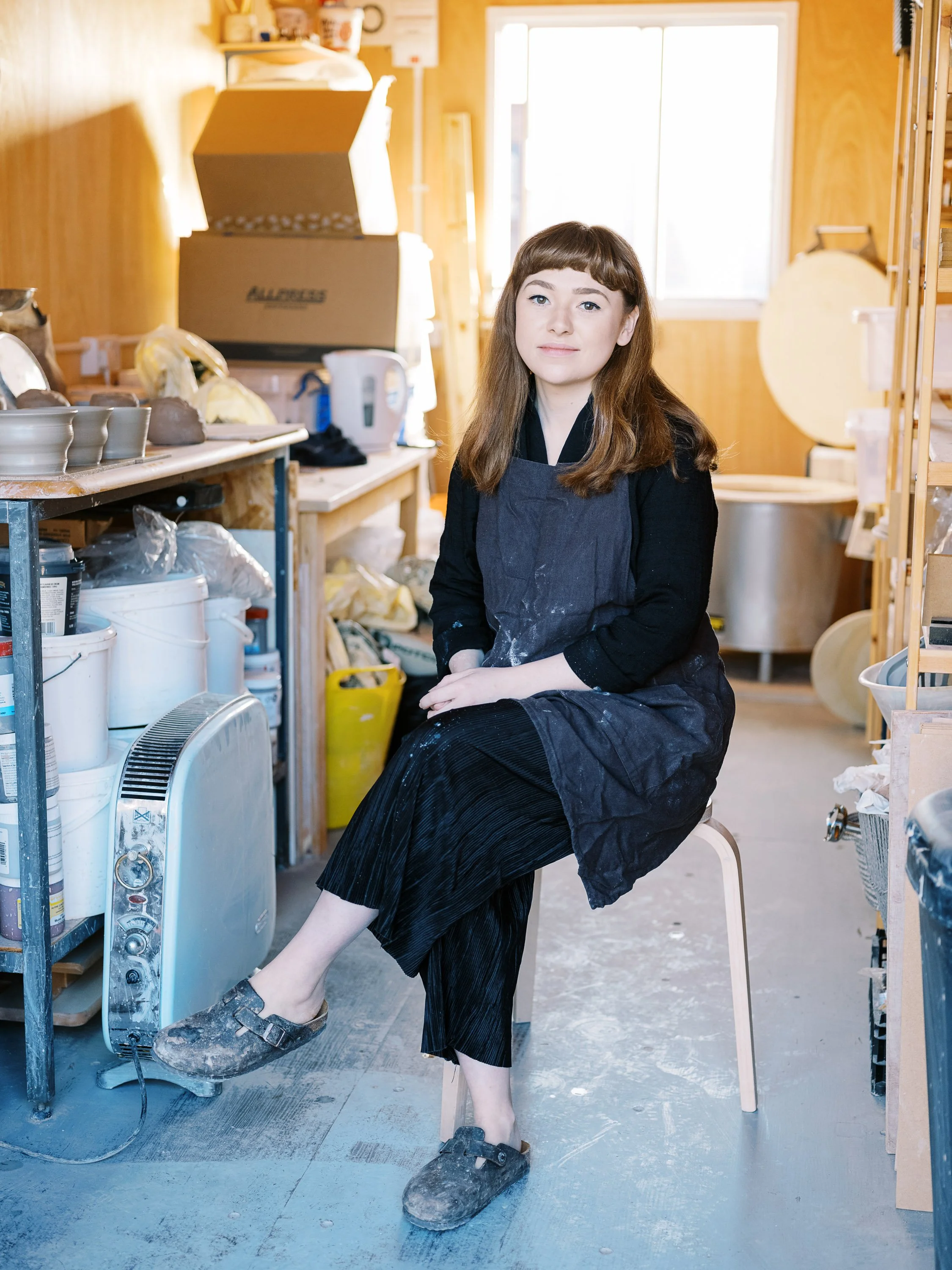 A woman with long brown hair and light skin, wearing a black shirt and apron, sitting on a stool in a pottery or ceramic studio. She has one leg crossed over the other, with her feet covered in clay. The studio has shelves with pottery supplies, a la