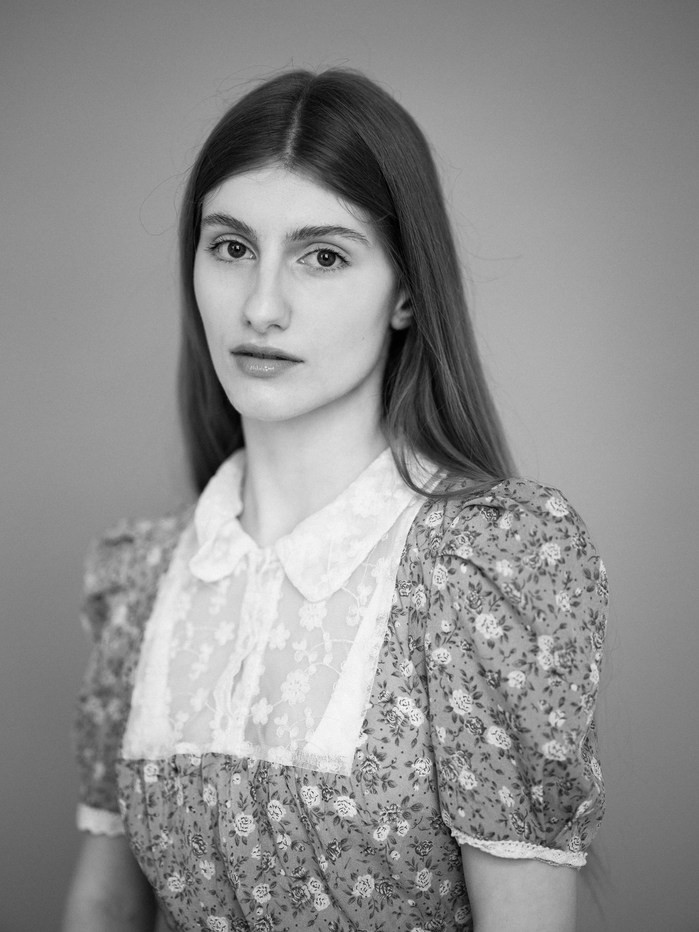 A black and white portrait of a young woman with long straight hair, wearing a floral dress with lace details, looking directly at the camera.