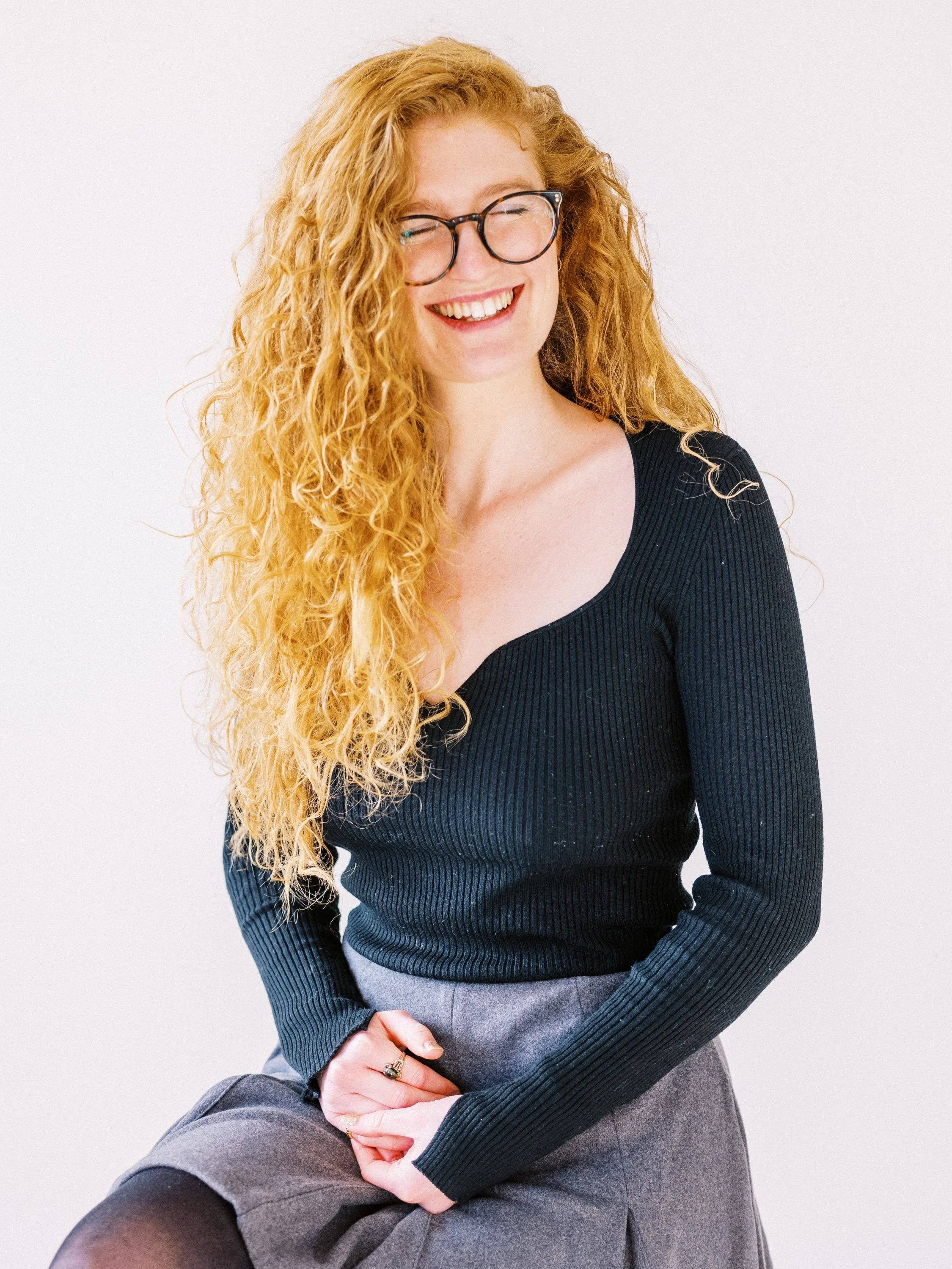 A woman with long, curly red hair and glasses, wearing a black ribbed sweater and a gray skirt, smiling with her eyes closed against a plain white background.