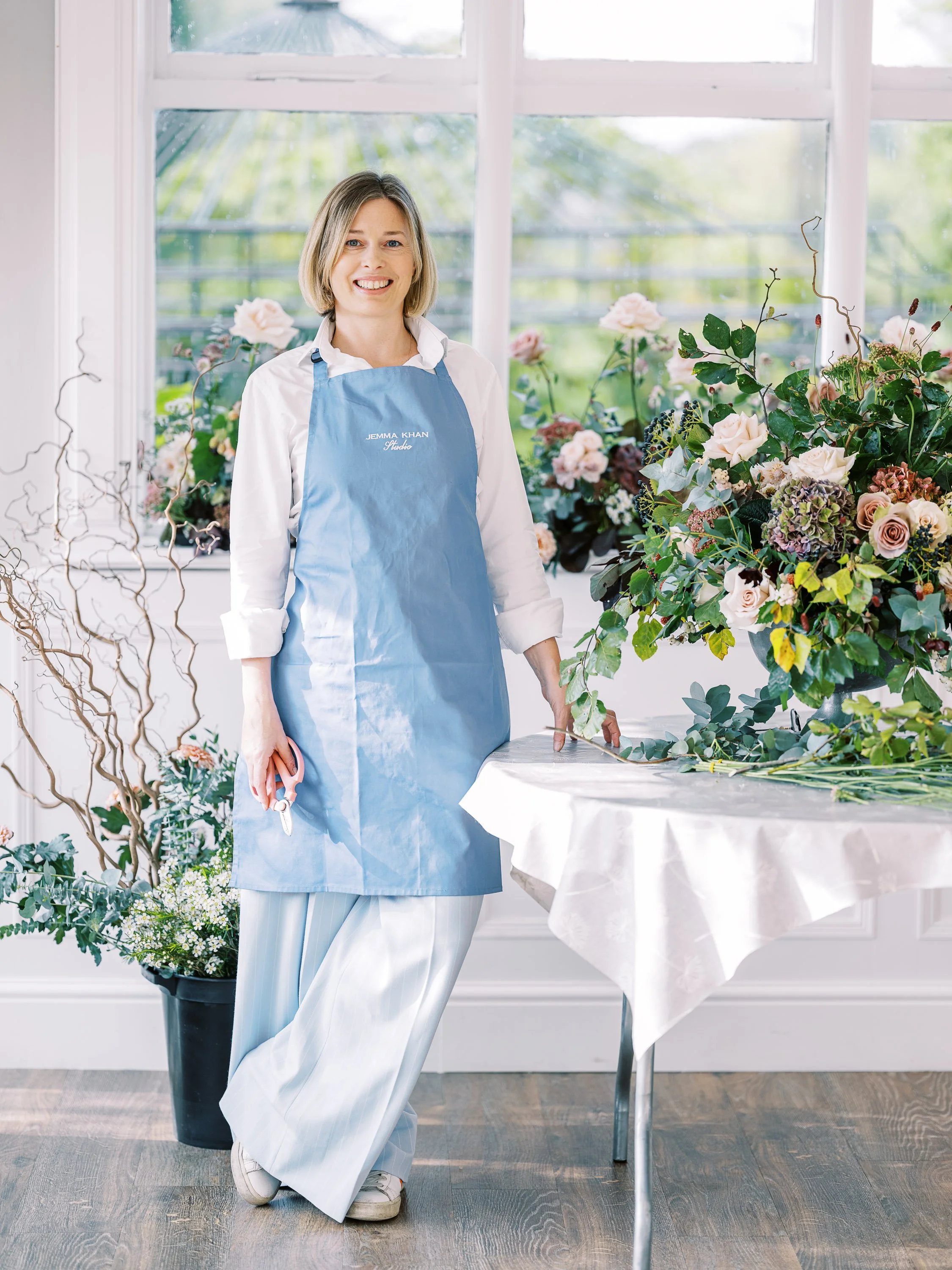 A woman in a blue apron standing next to a table with floral arrangements, in a bright room with large windows.