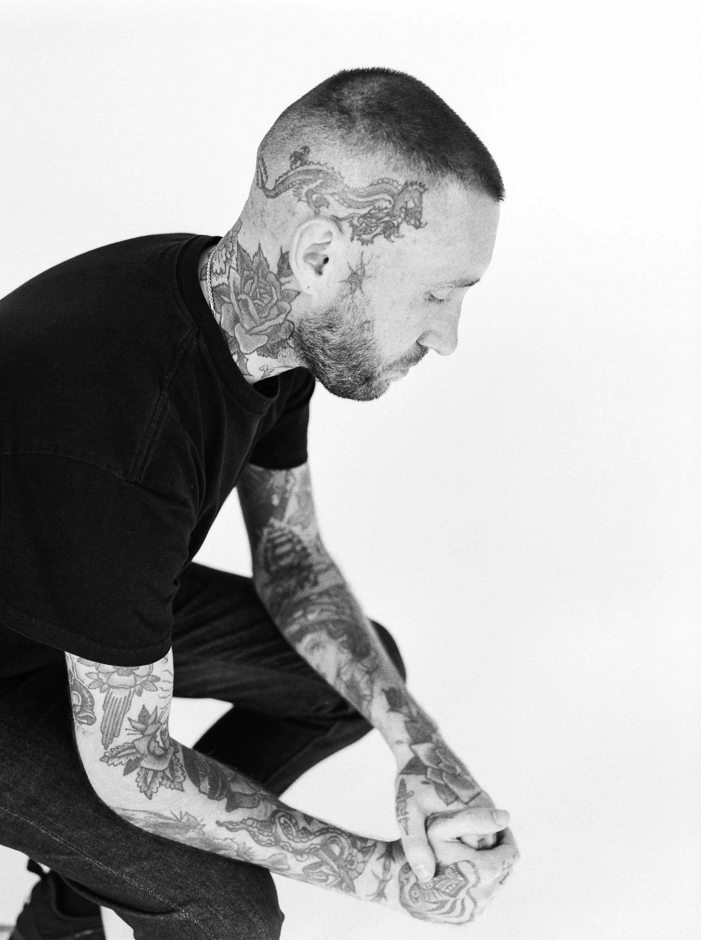 A man with tattoos on his head, neck, and arms, wearing a black t-shirt, sitting with his head bowed and eyes closed against a plain white background.