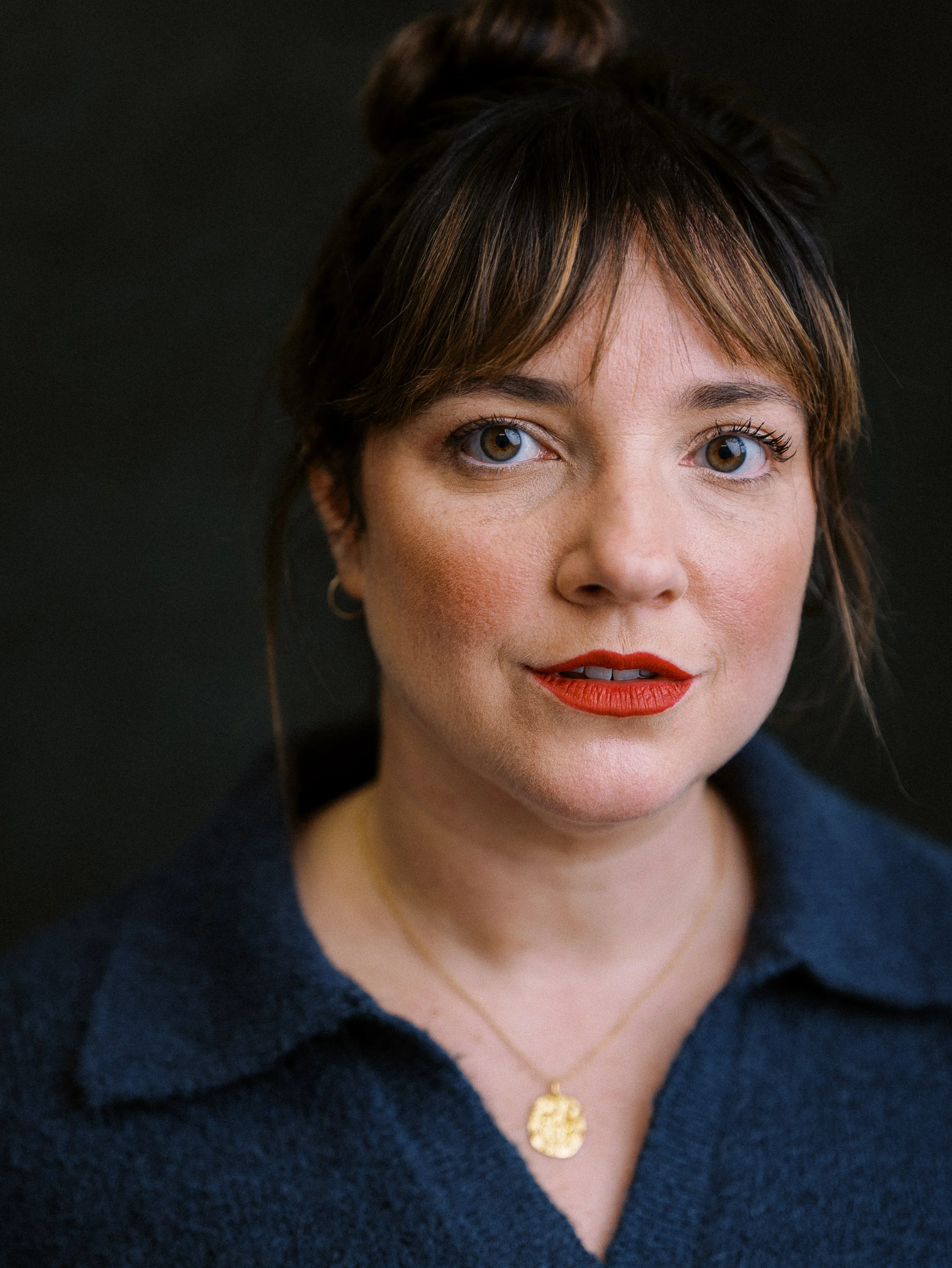 Close-up portrait of a woman with short brown hair, blue eyes, wearing red lipstick, gold earrings, a gold pendant necklace, and a navy blue collared top against a dark background.