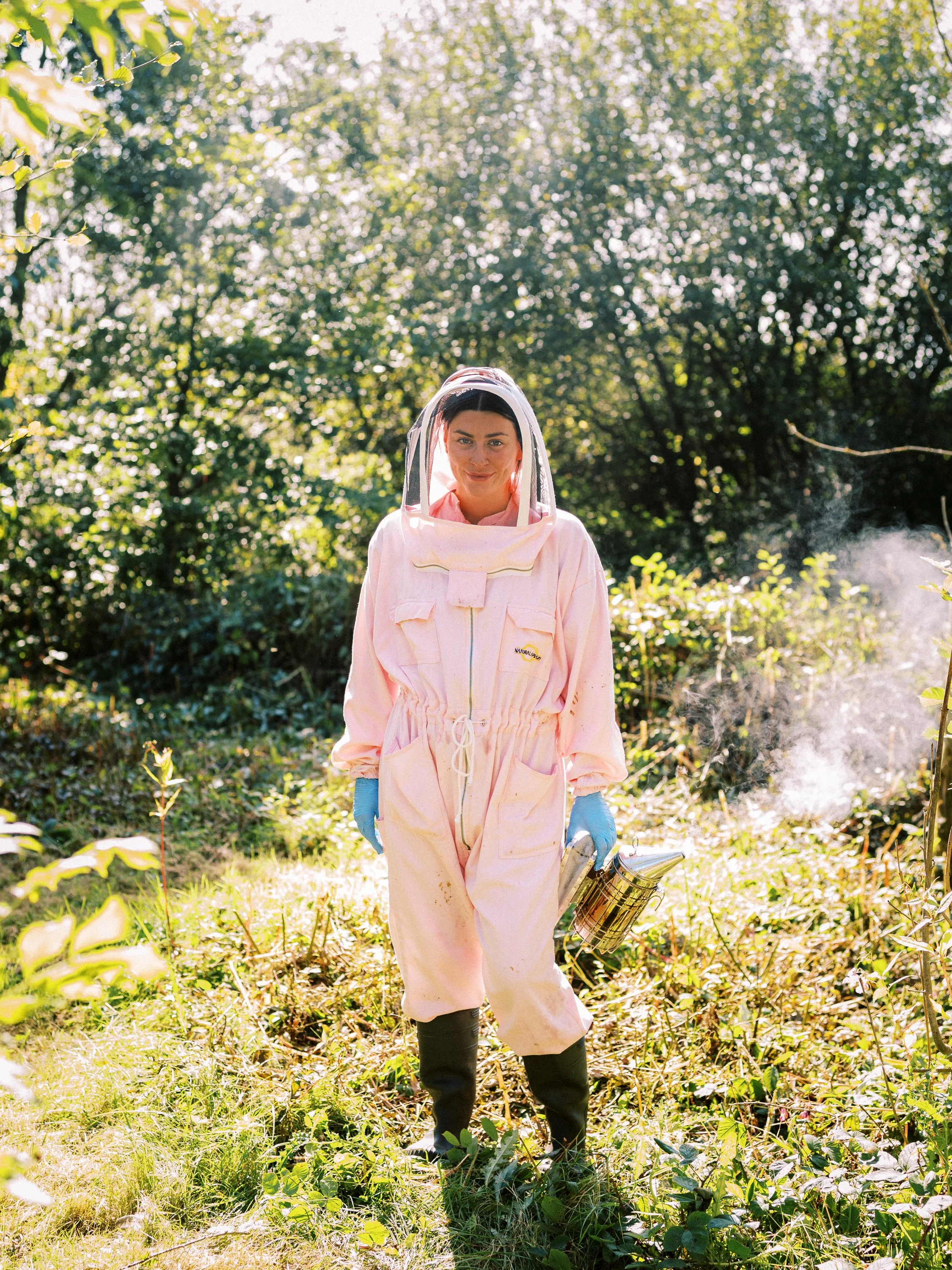 Woman wearing a pink protective suit and face shield, holding a spray canister, walking through a lush green outdoor area