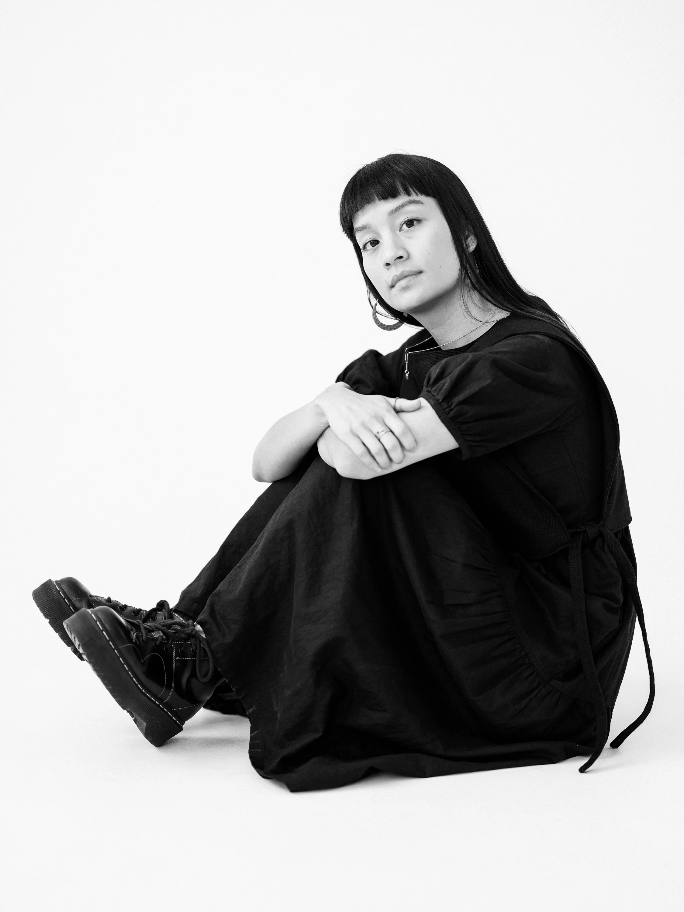A young woman with dark hair and bangs, sitting on the floor with her arms resting on her knees, dressed in black, against a plain white background.