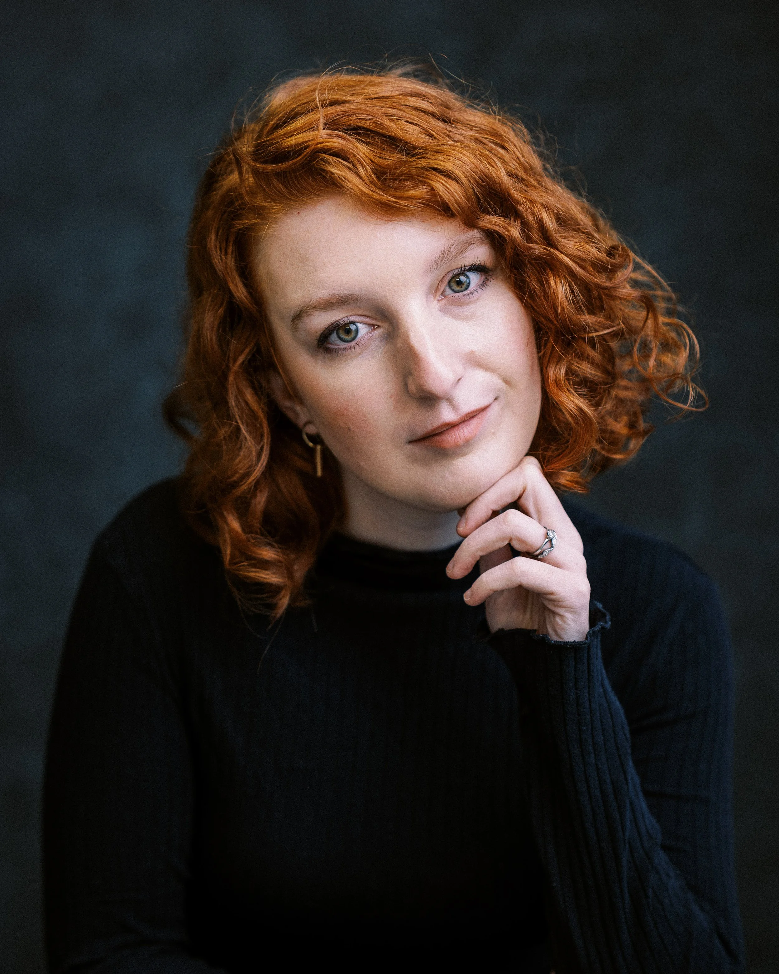 Portrait of a woman with curly red hair in a black top, resting her chin on her hand, against a dark background.
