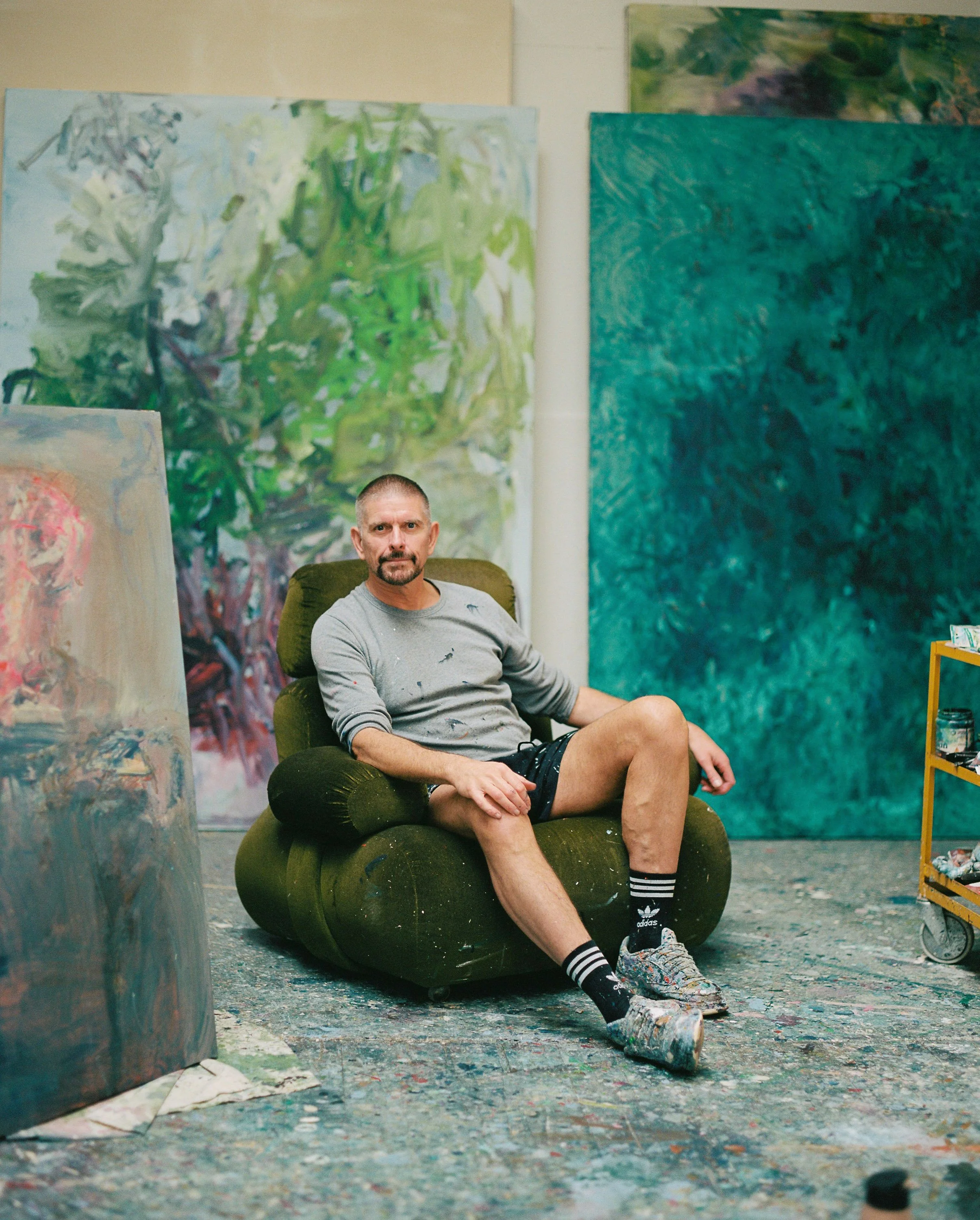 An artist sitting in a green armchair in an art studio surrounded by abstract paintings and a cart with art supplies.