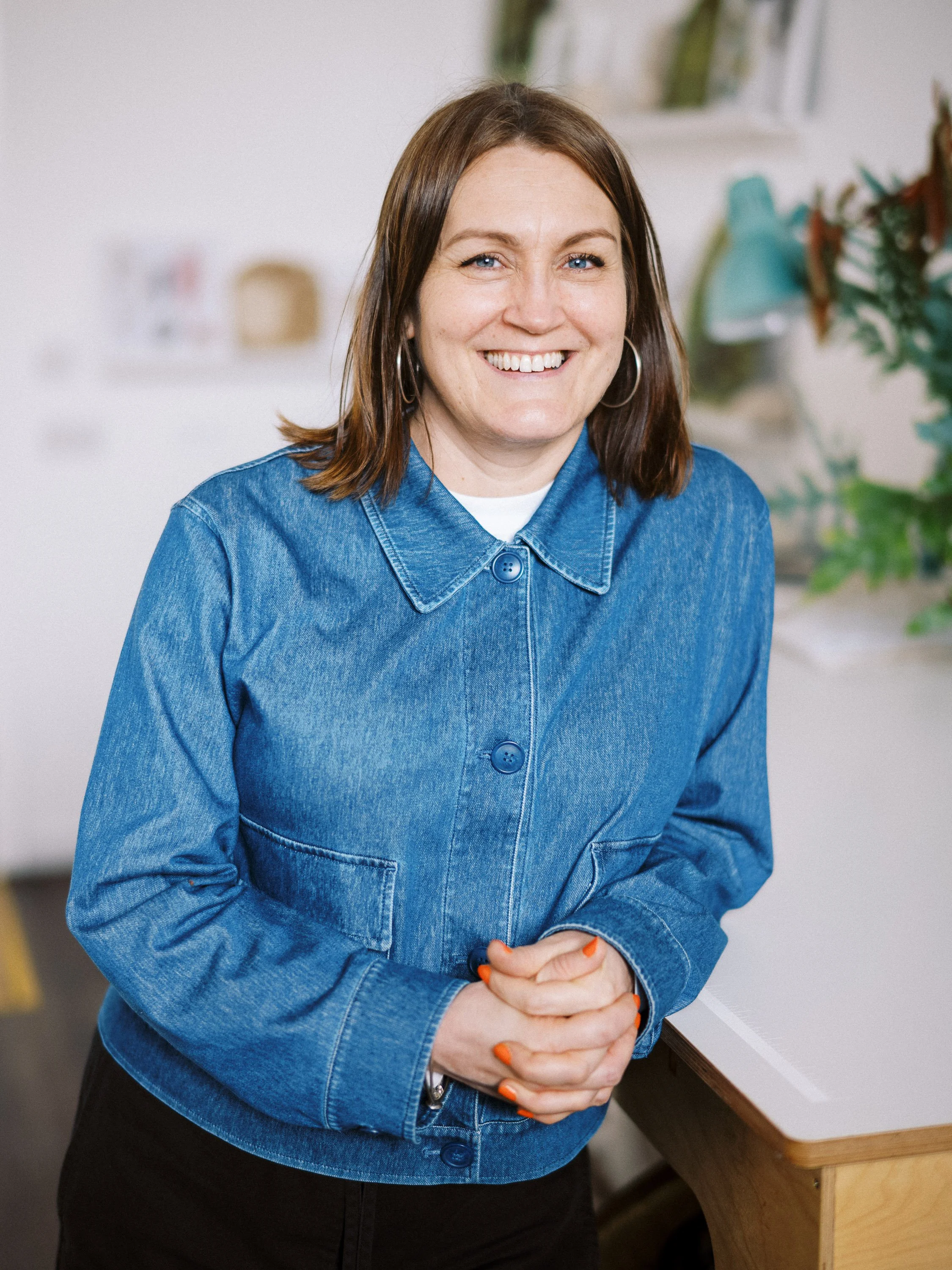 A woman with shoulder-length brown hair, blue eyes, and hoop earrings, smiling and standing indoors with hands clasped. She is wearing a blue denim jacket over a white top.