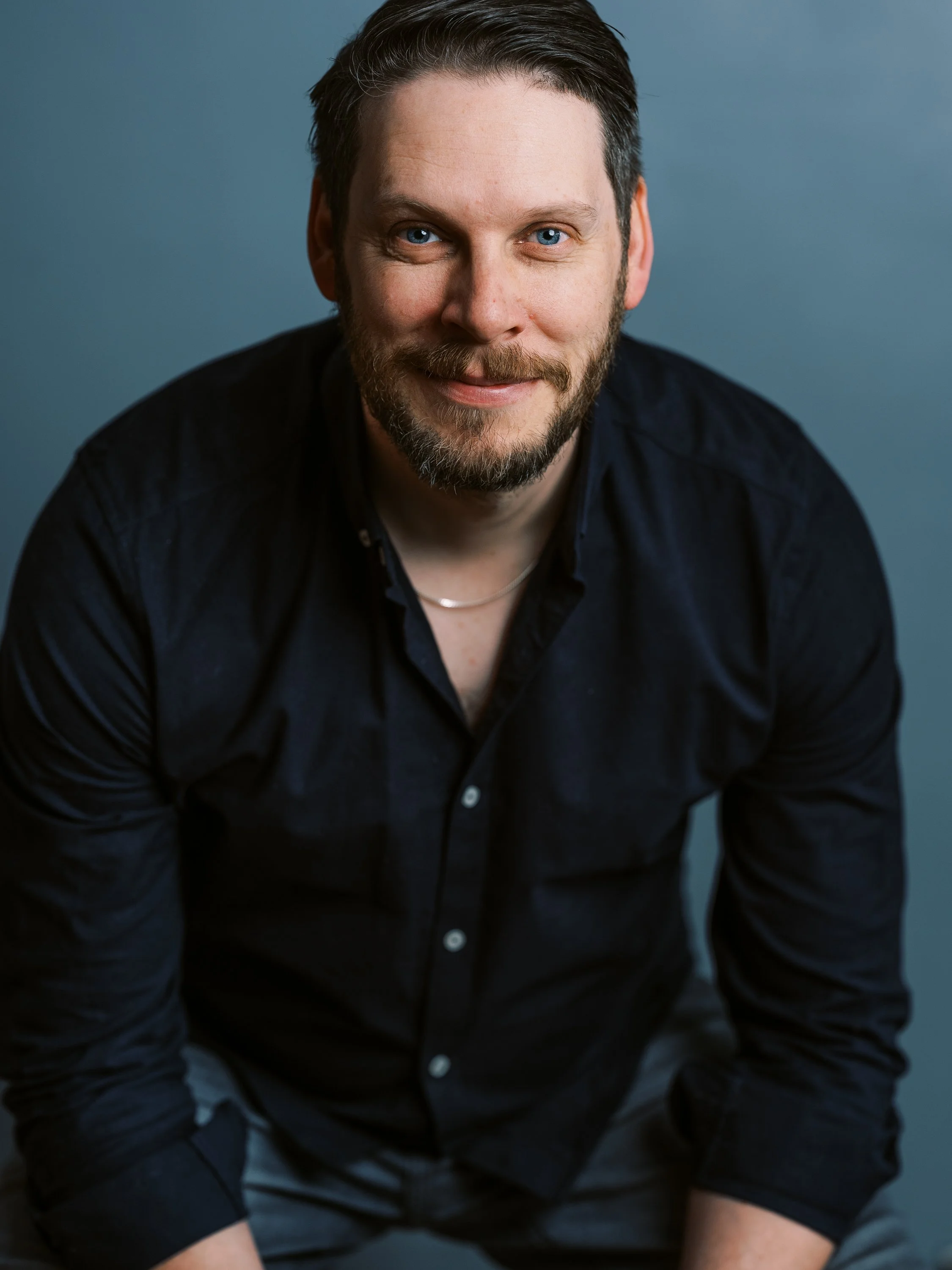 A man with light skin, blue eyes, dark hair, and a beard, wearing a black shirt, smiling at the camera against a blue background.