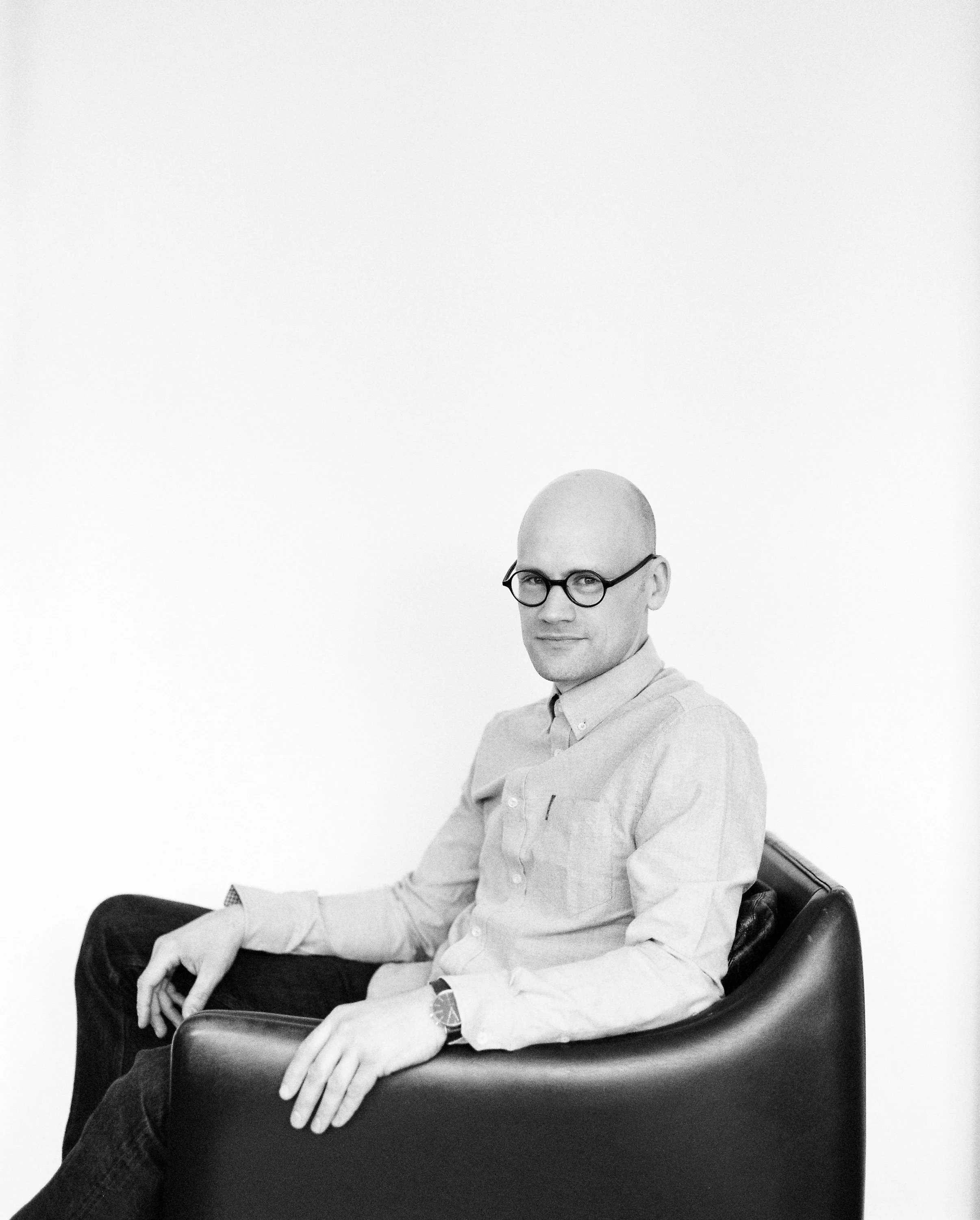 Black and white photo of a bald man wearing glasses, a light-colored button-up shirt, sitting in a modern armchair against a plain white background.