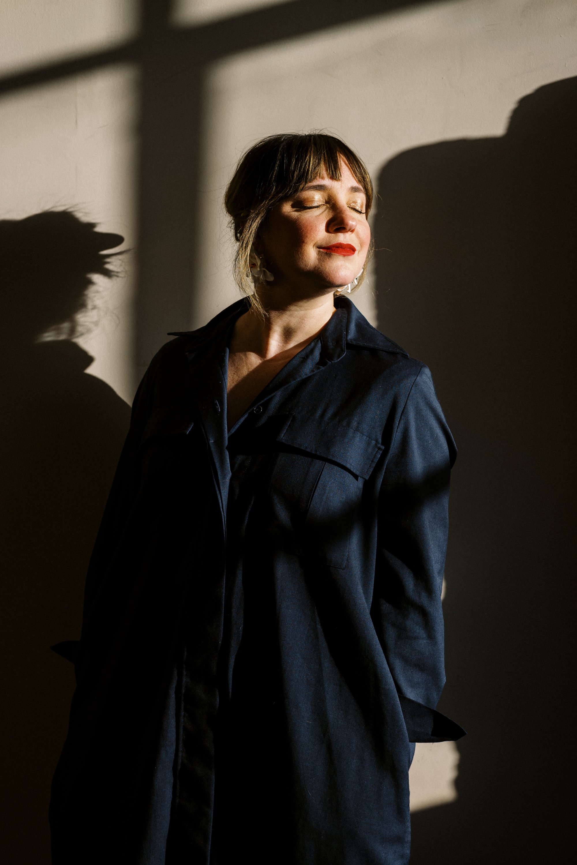 A woman with short hair and red lipstick standing with her eyes closed, smiling slightly, in front of a wall with shadows cast by sunlight.