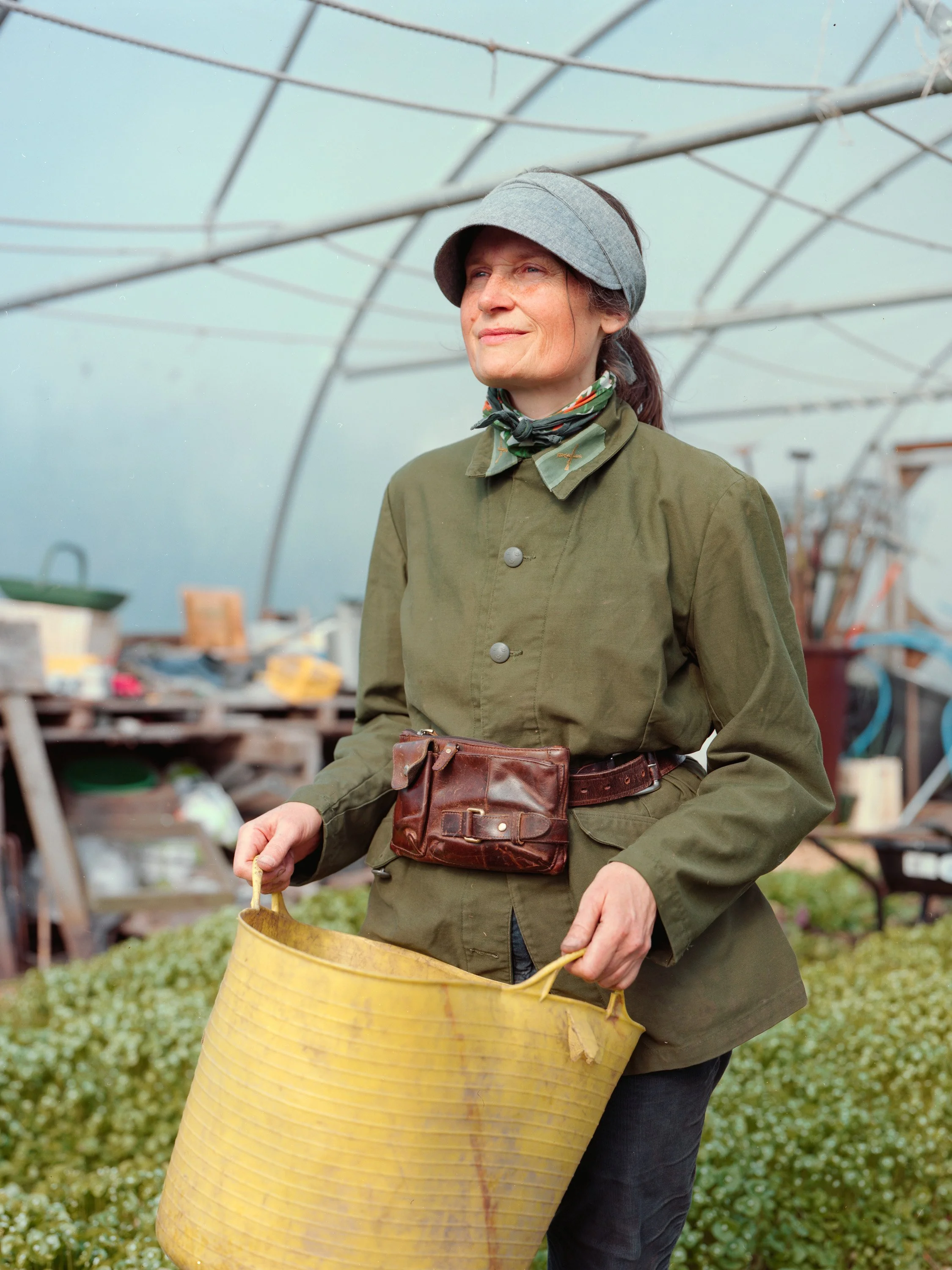 A woman in a green jacket, gray visor, and patterned scarf stands in a greenhouse, holding a yellow container, with plants around her.