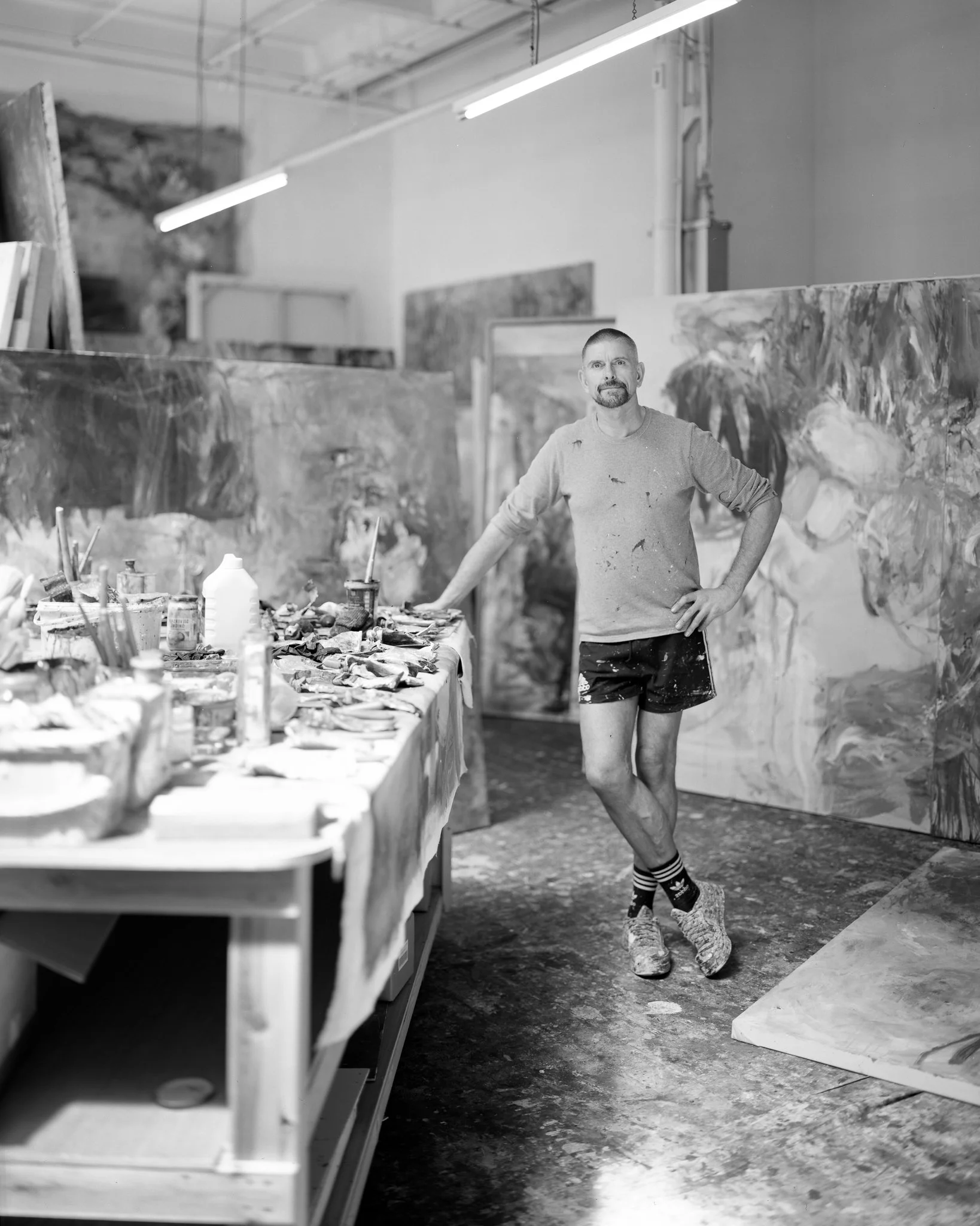 An artist stands in his studio, surrounded by large abstract paintings, with a cluttered table of art supplies. He is casually dressed, wearing a long-sleeve shirt, shorts, and patterned socks, with paint splatters on his clothes.