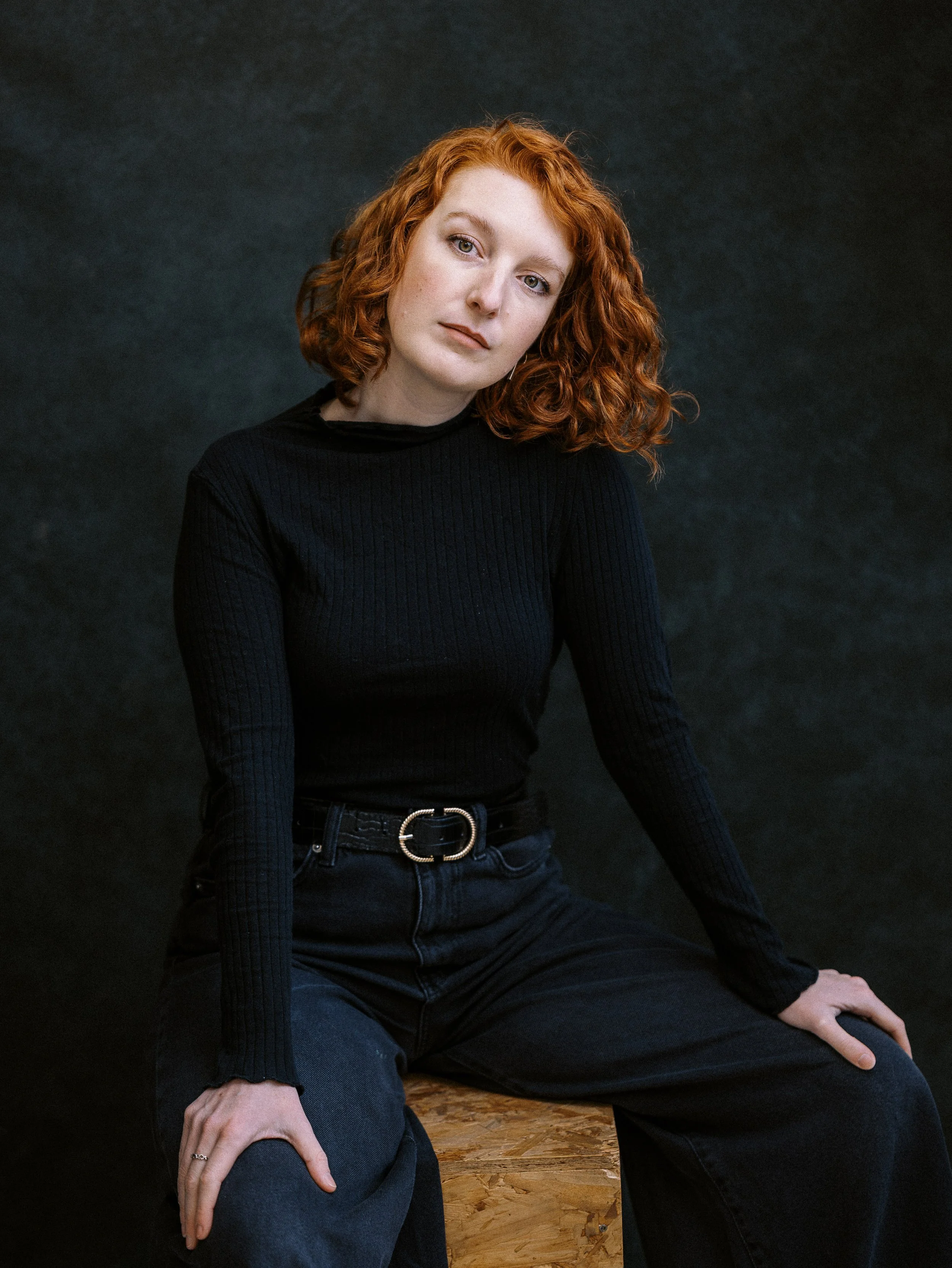 A woman with curly red hair wearing a black ribbed long sleeve shirt and black pants, sitting on a wooden box against a dark background.