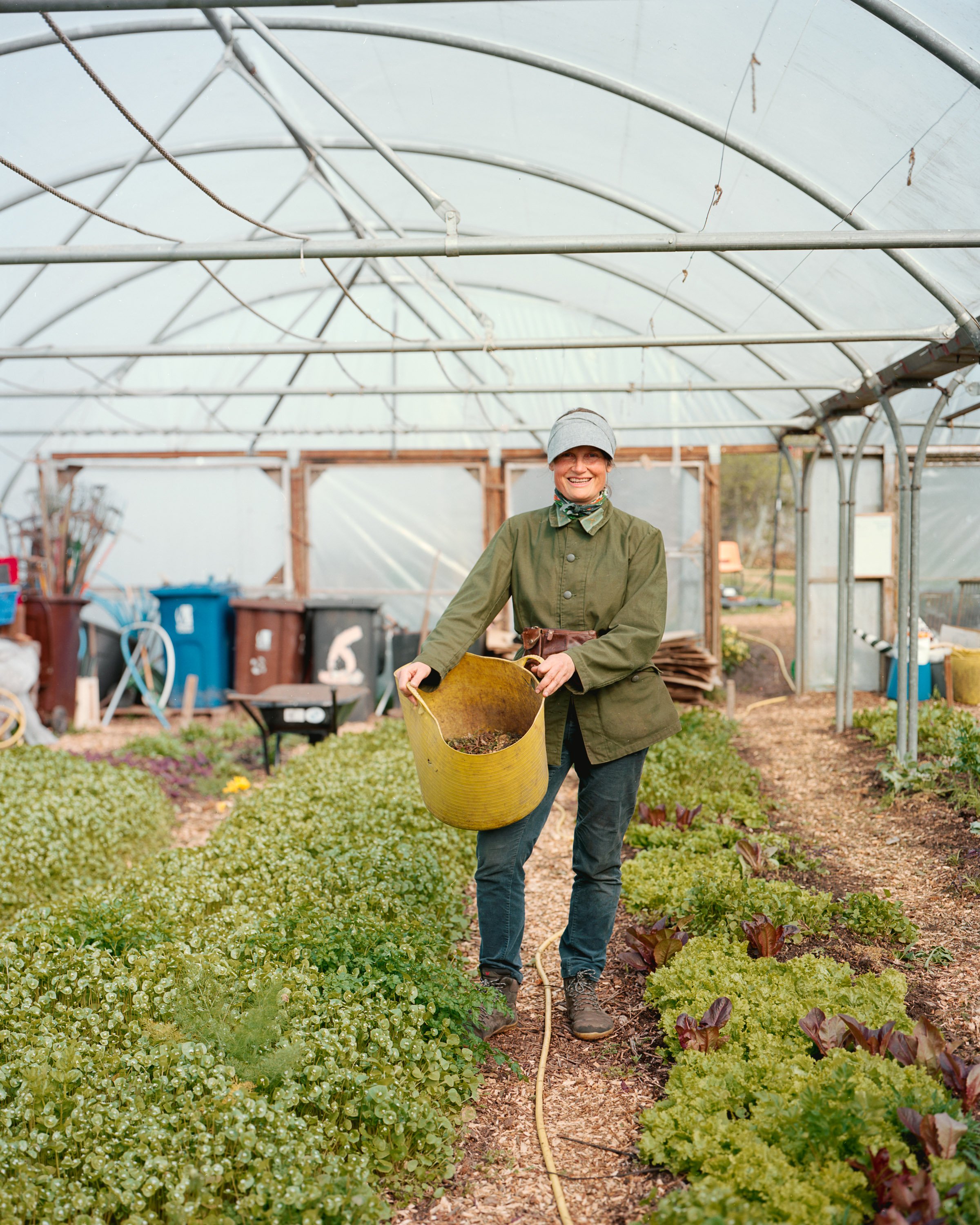 A smiling woman in gardening attire holding a yellow bucket inside a greenhouse, surrounded by greenery and rows of leafy plants.
