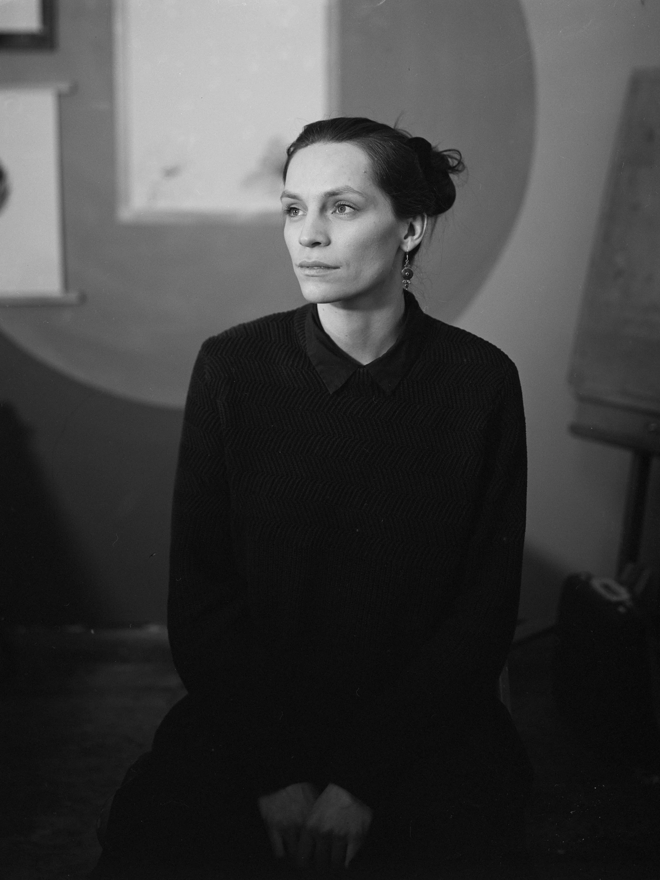 A black and white photo of a woman with dark hair styled in an updo, sitting and looking to her left, wearing a dark sweater with a collared shirt underneath and dangling earrings.