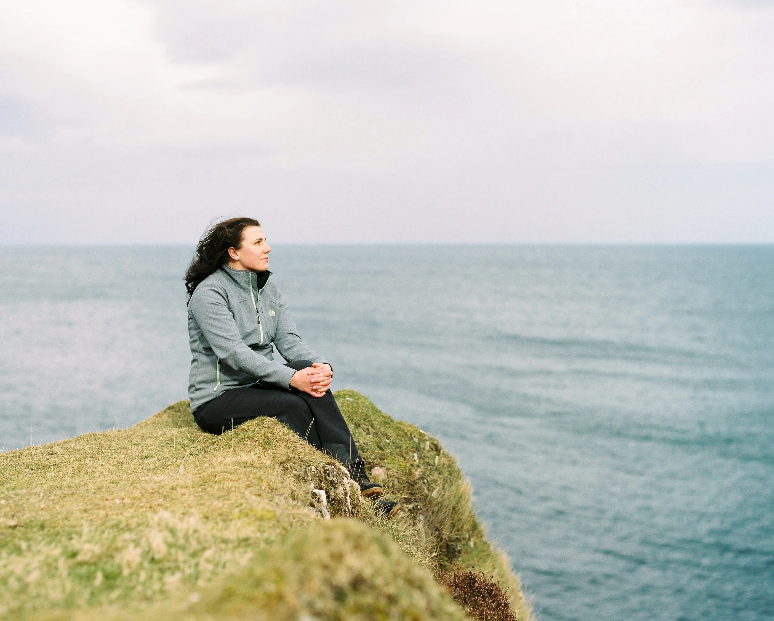 A woman sitting on a grassy cliff overlooking the ocean, wearing a gray jacket and black pants, gazing contemplatively at the water.