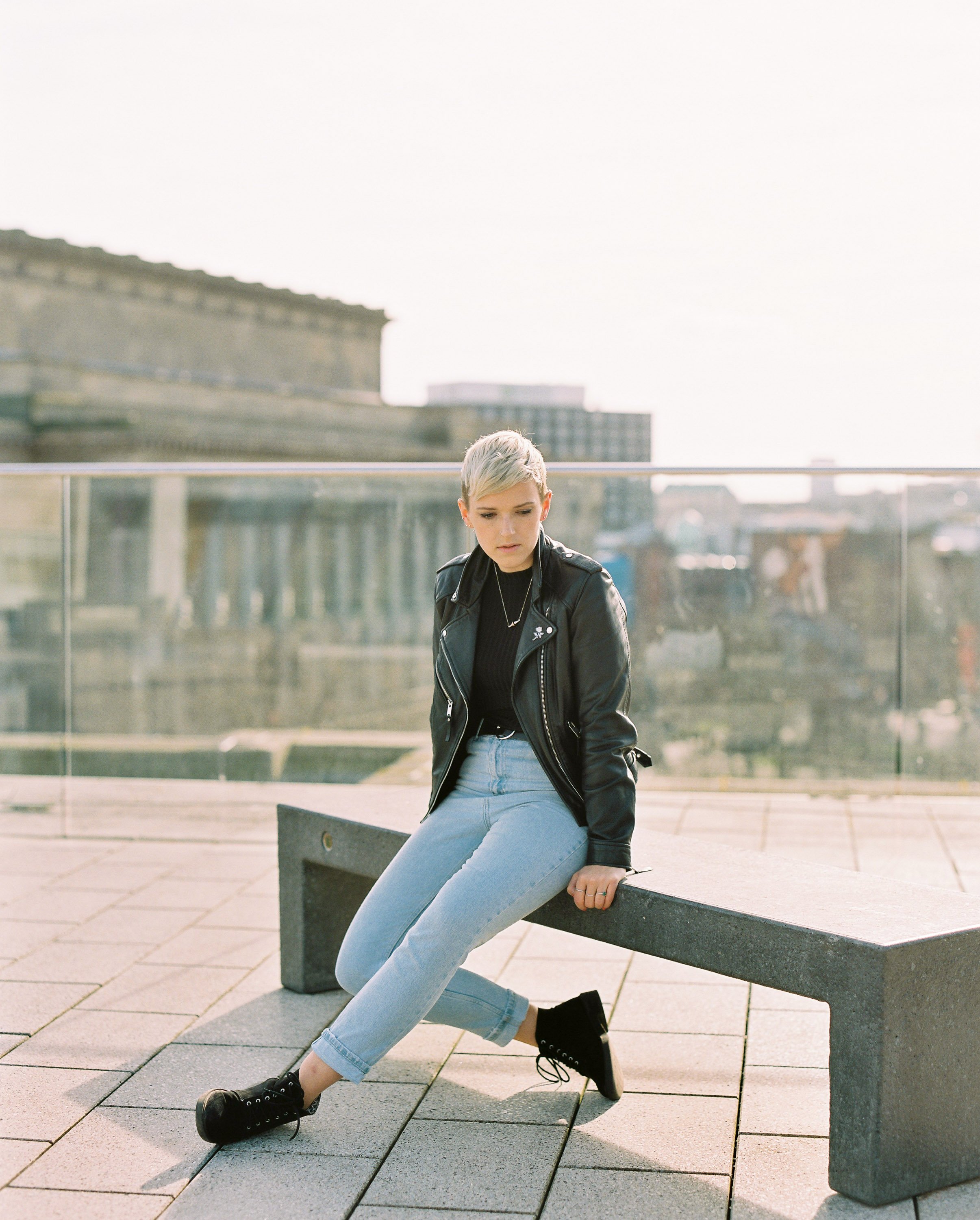 A young woman with short blonde hair sitting on a concrete bench outdoors, wearing a black leather jacket, black top, light blue jeans, and black sneakers.