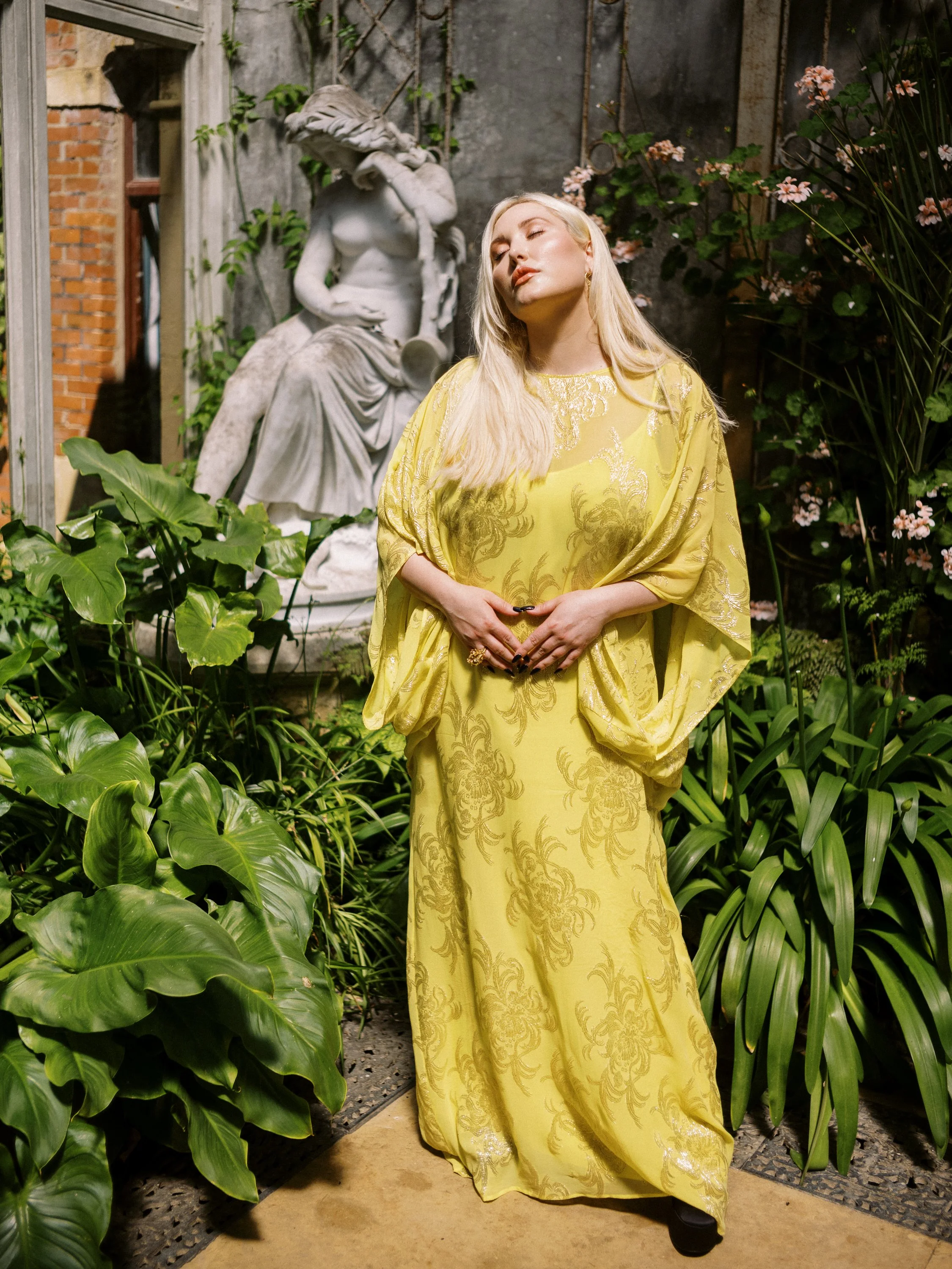 A woman with long blonde hair standing in a garden with lush green plants and pink flowers, wearing a flowing yellow dress with intricate patterns, with eyes closed and hands resting on her stomach, in front of a classical sculpture of a seated woman