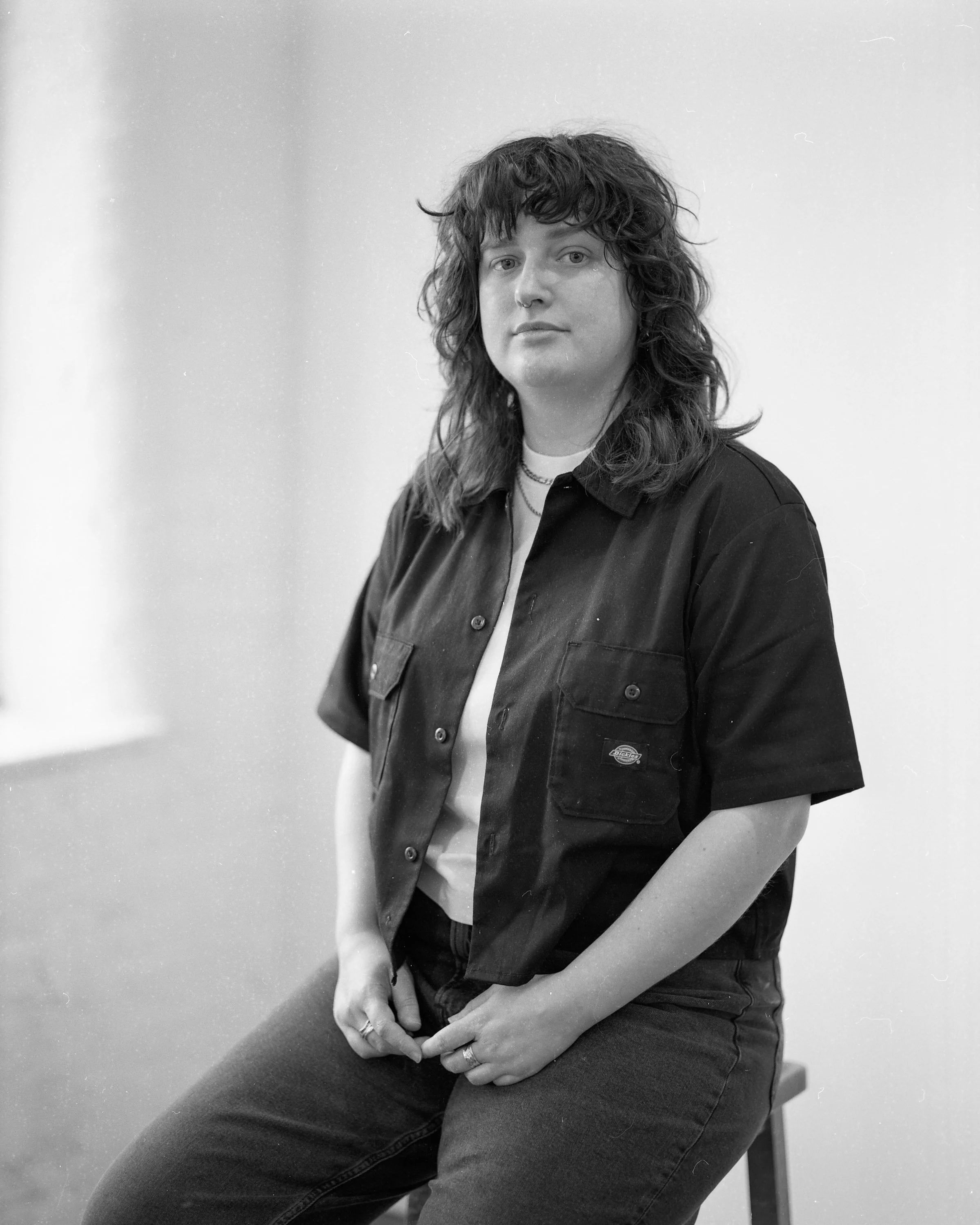 Black and white photo of a woman with long, wavy hair sitting on a stool against a plain background. She wears a short-sleeved button-up shirt, rings, and a necklace, looking directly at the camera.