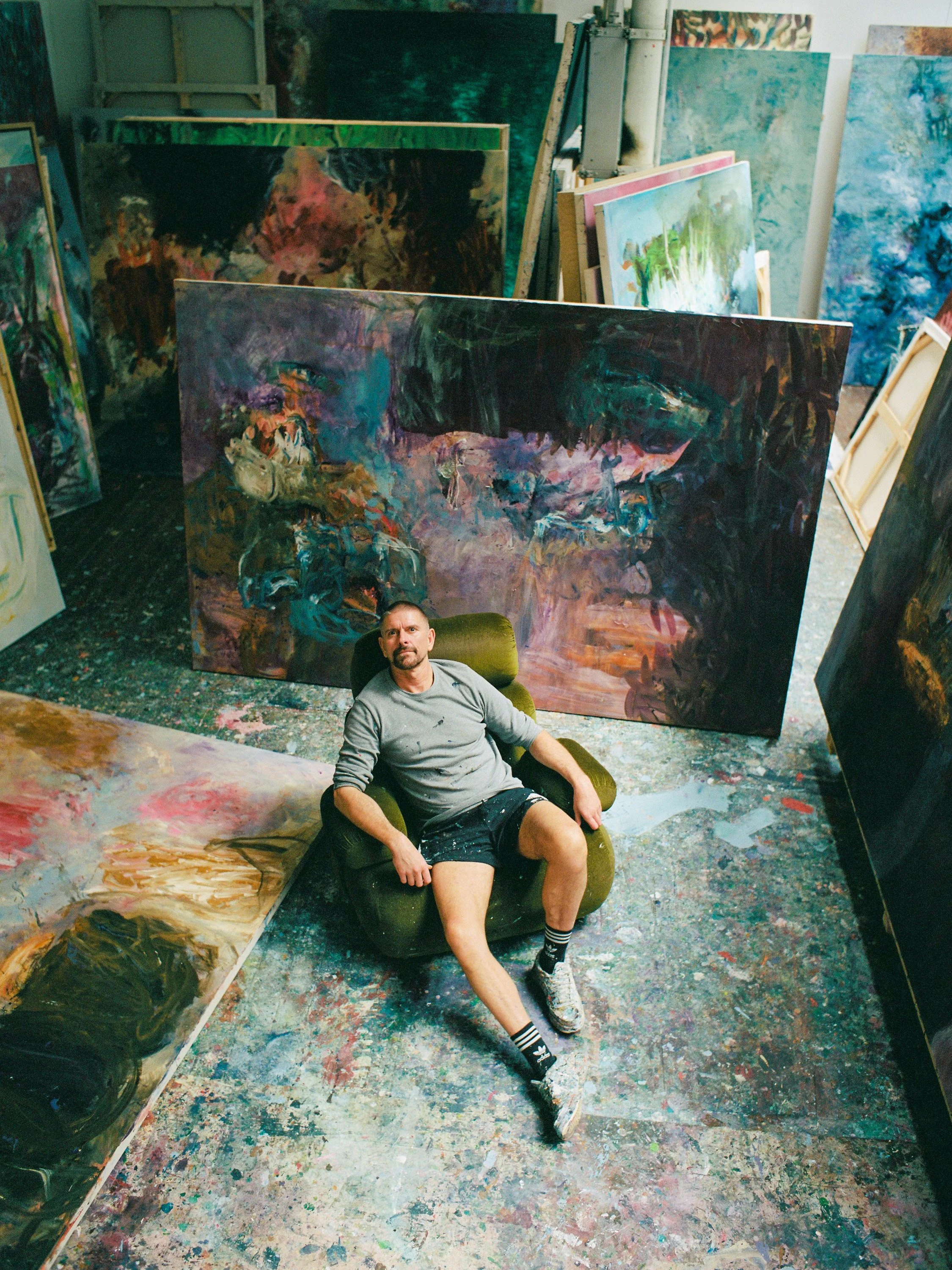 An artist sits in a green armchair in a cluttered art studio surrounded by large abstract paintings on the floor and leaning against the walls, with a colorful, paint-splattered floor.