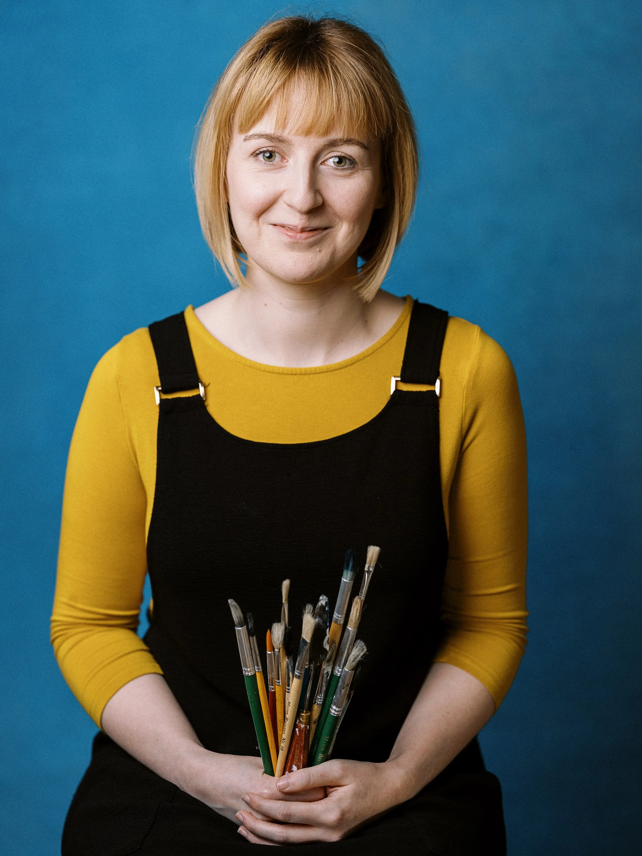 A woman with short red hair and a yellow long-sleeve shirt, wearing a black apron, holding a bunch of paintbrushes, standing against a blue background.
