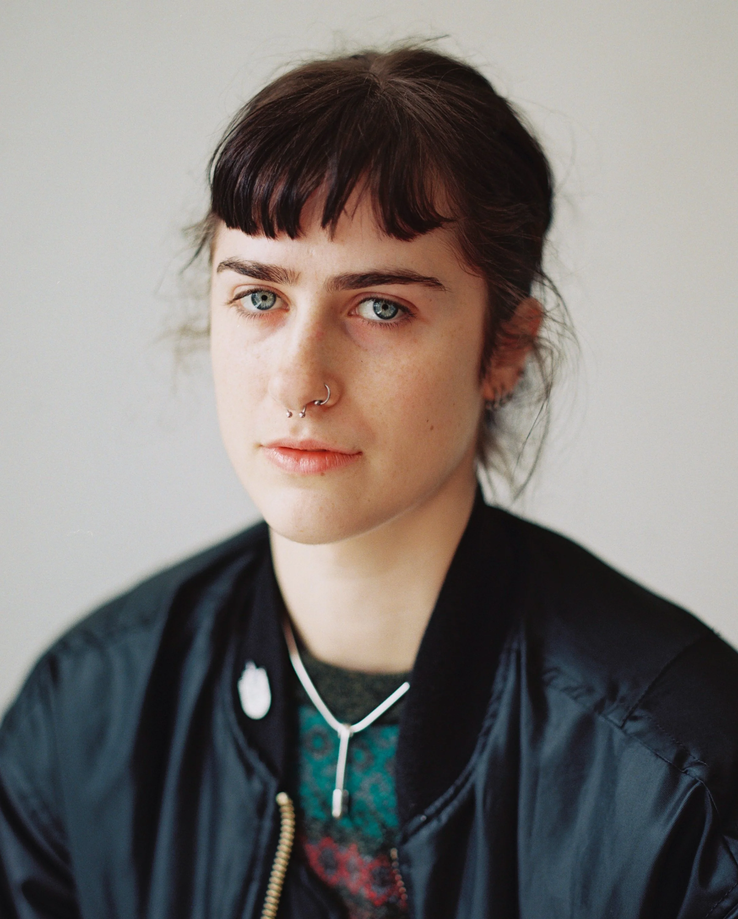 Close-up portrait of a young woman with short dark hair, blue eyes, and pierced nose, wearing a black jacket and necklace, against a plain light background.