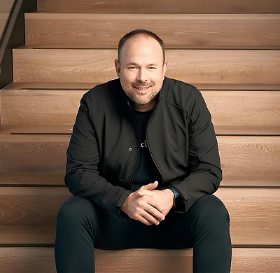 A man sitting on wooden stairs, smiling, wearing a black jacket and black pants.