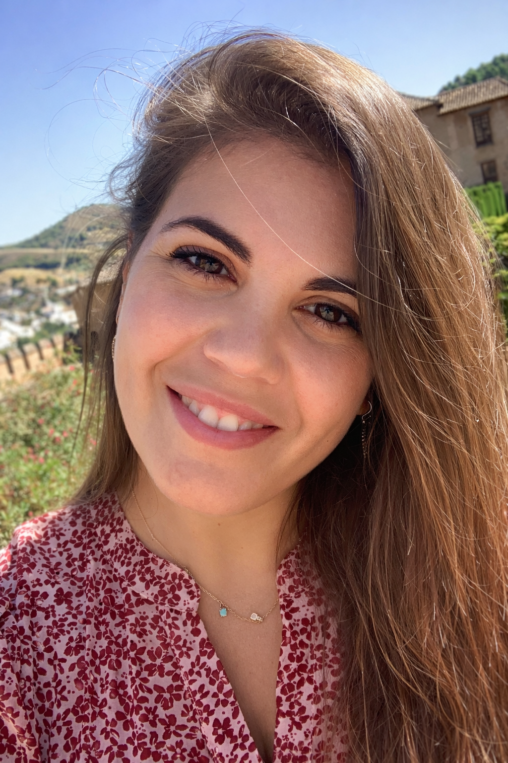 Retrato de una mujer joven con cabello largo y rizado, sonriendo en un día soleado con paisaje de colinas y edificios antiguos de fondo.