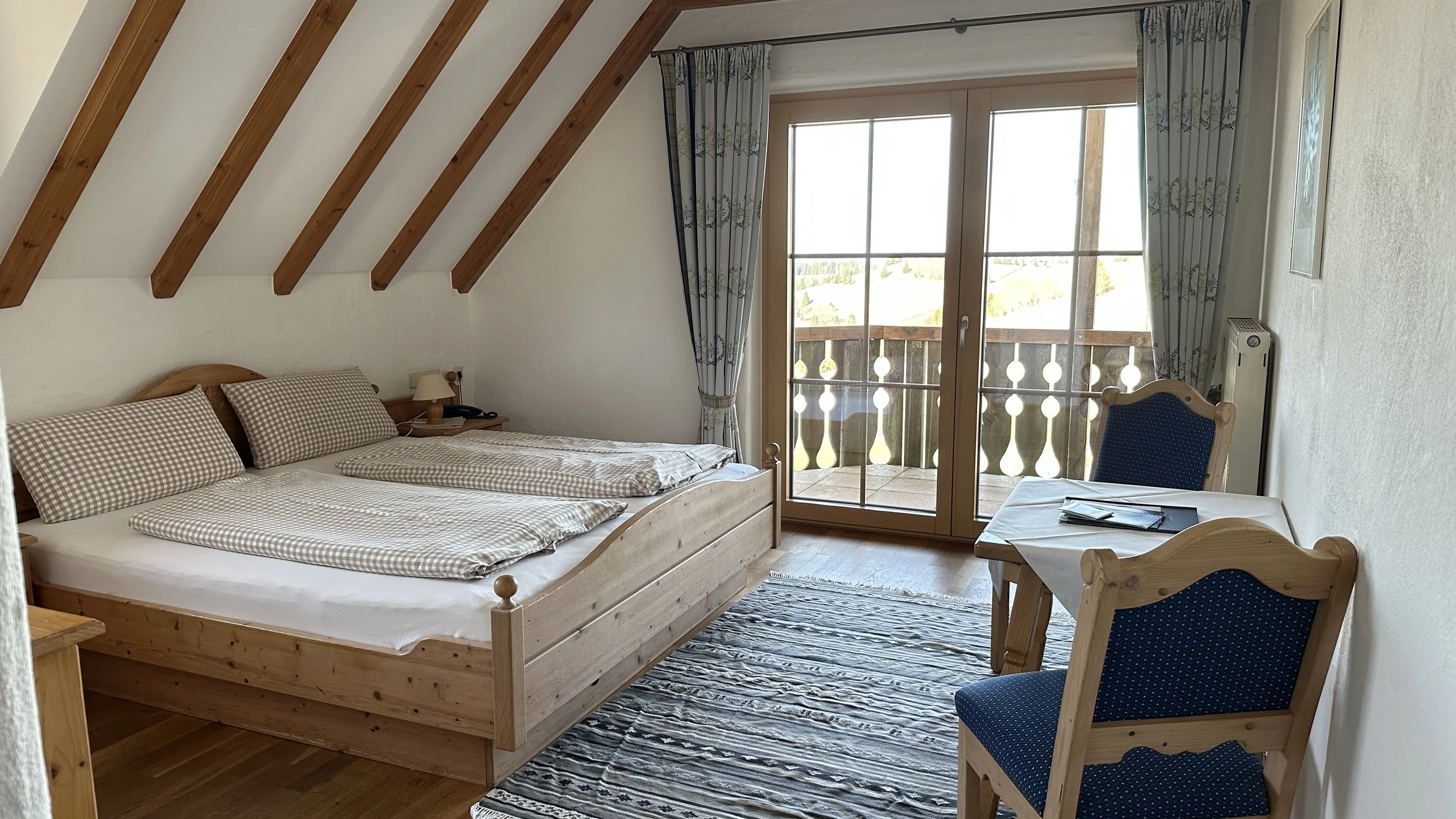 Ein gemütliches Hotelzimmer mit Holzdielenboden, einem Doppelbett, einem kleinen Tisch mit zwei blauen Polsterstühlen und einem Balkon mit Blick ins Grüne.