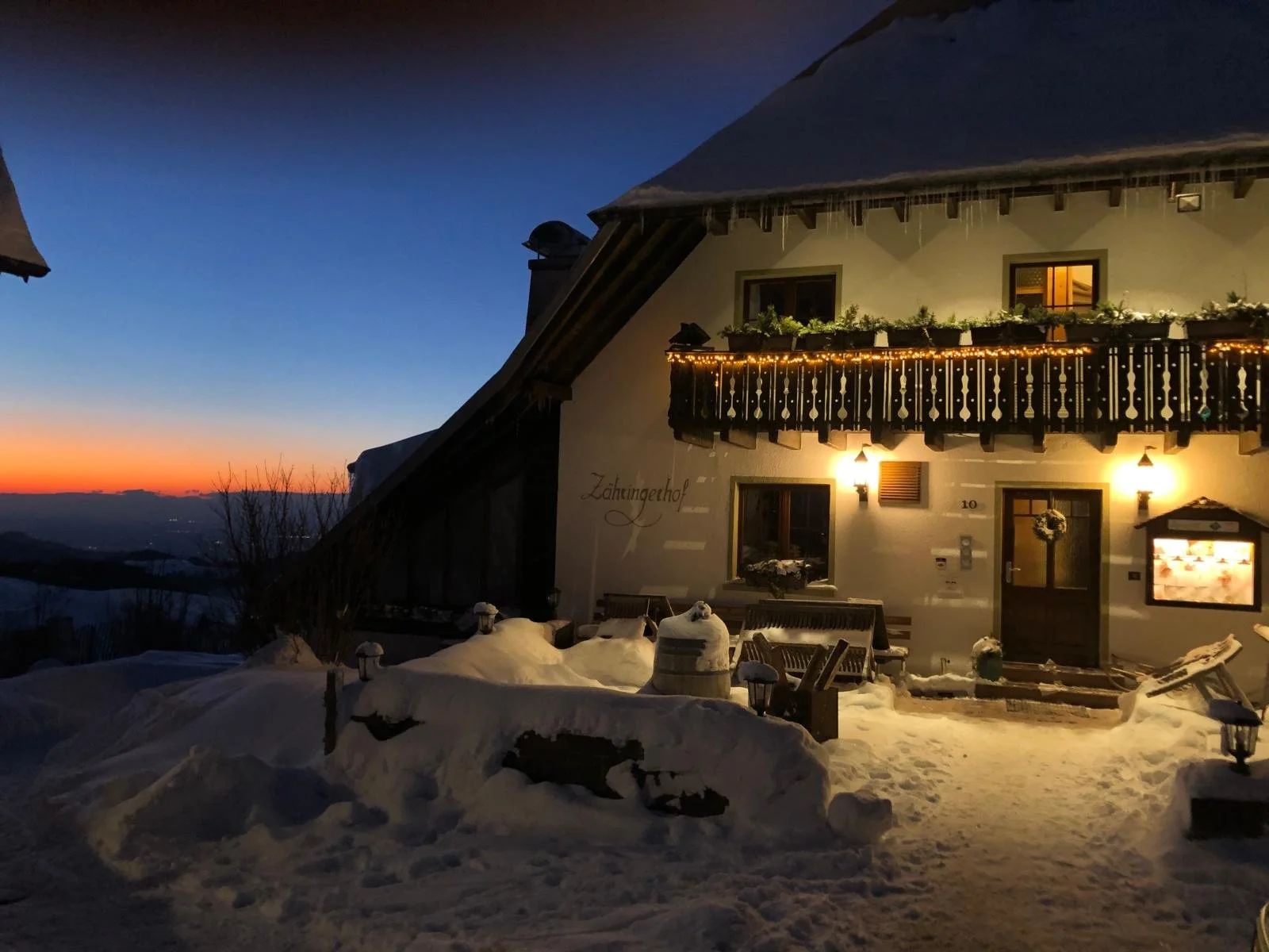 Ein verschneites Haus bei Sonnenuntergang mit warmem Licht aus den Fenstern und einem Balkon mit Girlanden. Der Himmel ist abends dunkel, die Landschaft im Hintergrund ist schneebedeckt.