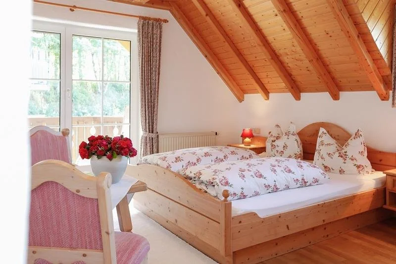 Ein Schlafzimmer mit Holzbalken an der Decke, Doppelbett mit floralbezogenen Kissen und Bettdecke, Nachttisch mit roter Lampe, großer Fenstertür mit Vorhängen, Balkon mit Blick auf grüne Bäume, Vase mit roten Blumen auf einem Tisch.