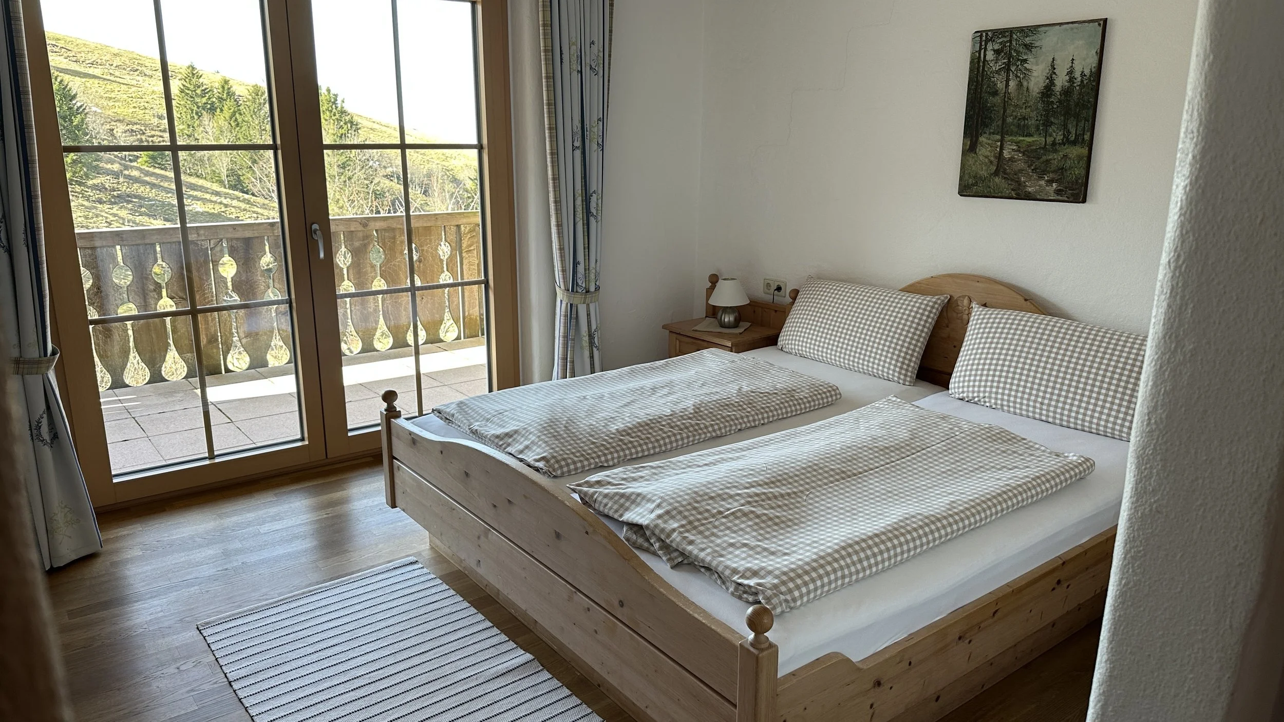 Ein Schlafzimmer mit Doppelbett, Holzmöbeln, wisch-gecheckter Bettwäsche und Zugang zu einem Balkon mit Blick auf eine grüne, hügelige Landschaft.