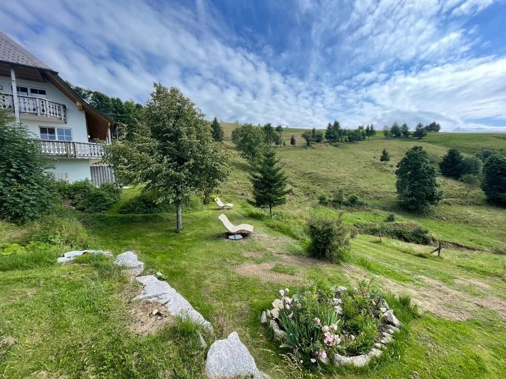 Grüner Garten mit Baum, Sträuchern, Blumen und Liegestühlen vor einem Haus auf einer Hügellandschaft unter einem bewölkten Himmel.