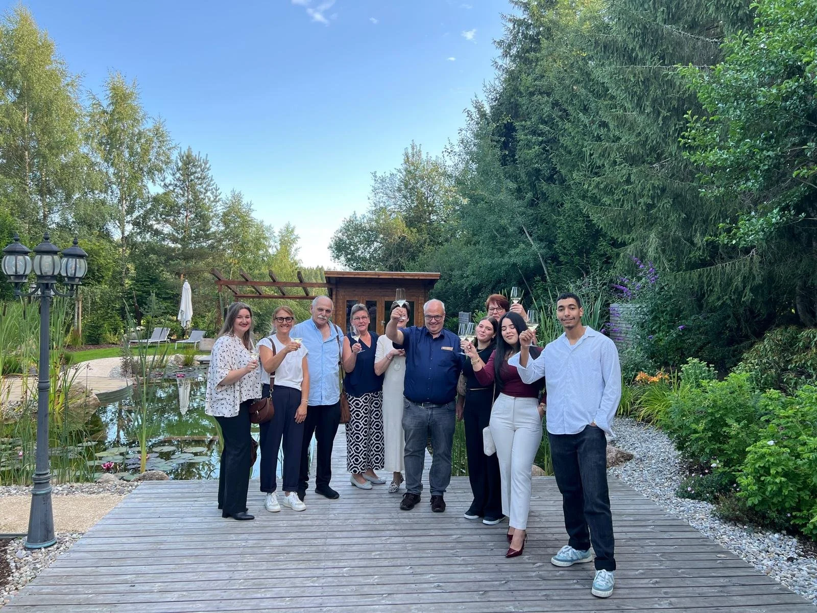 Eine Gruppe von Menschen, die im Garten bei Sonnenuntergang anstoßen, stehen auf einem Holzsteg neben einem Teich mit Seerosen, umgeben von Bäumen und Pflanzen.