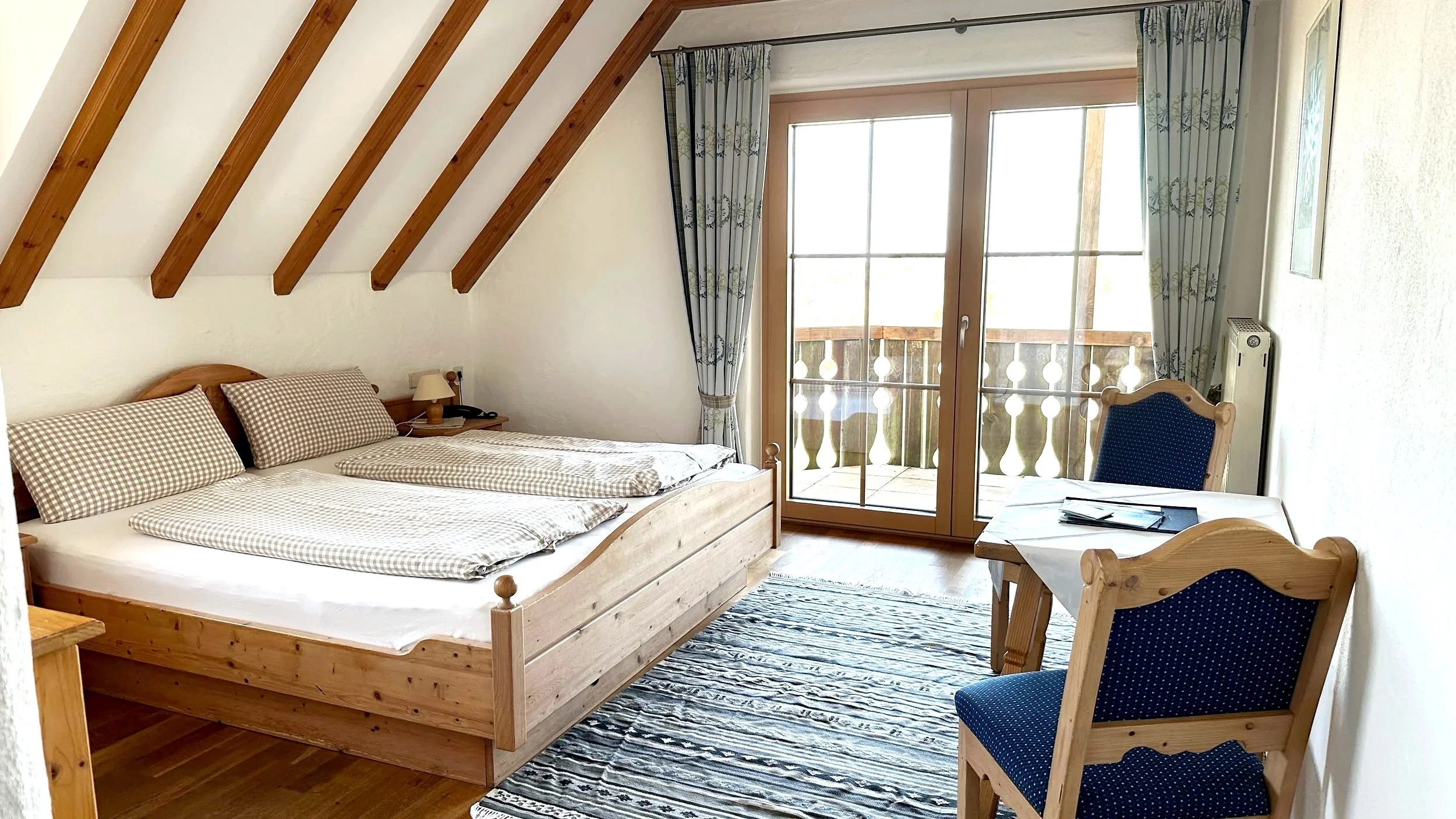 Ein Schlafzimmer mit einem Doppelbett, eines kleinen Tisches mit einem Telefon, einer Leselampe, einem Stuhl mit dunkelblauem Bezug, großen Fenstern und einer Tür zu einem Balkon, in einem Raum mit Holzdecke und weißen Wänden.