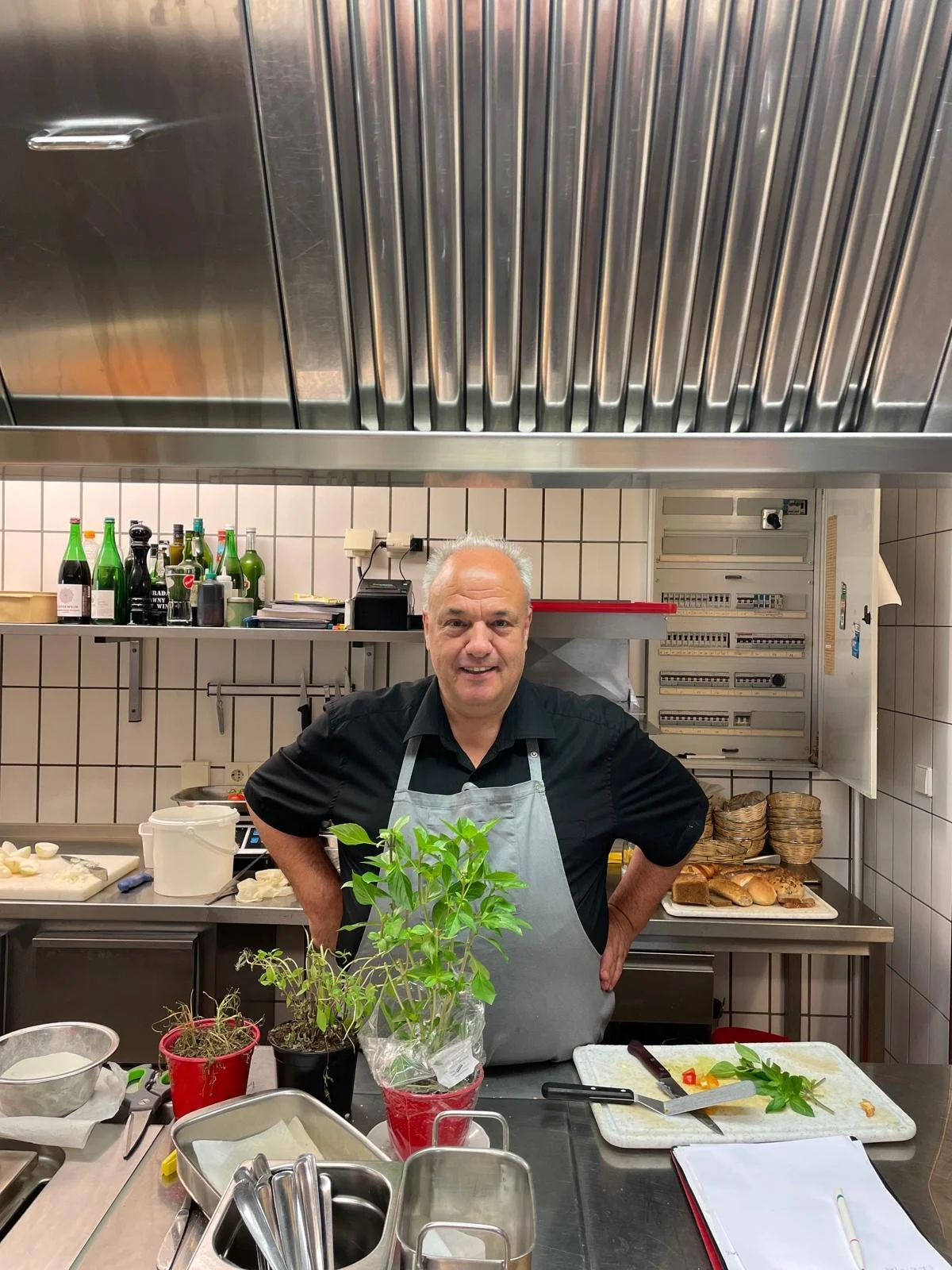 Ein lachender Mann in einer schwarzen Schürze steht in einer Küchenküche vor einem mit Kräutern und Küchenutensilien bedeckten Tresen, im Hintergrund sind Getränke und Backwaren sichtbar.